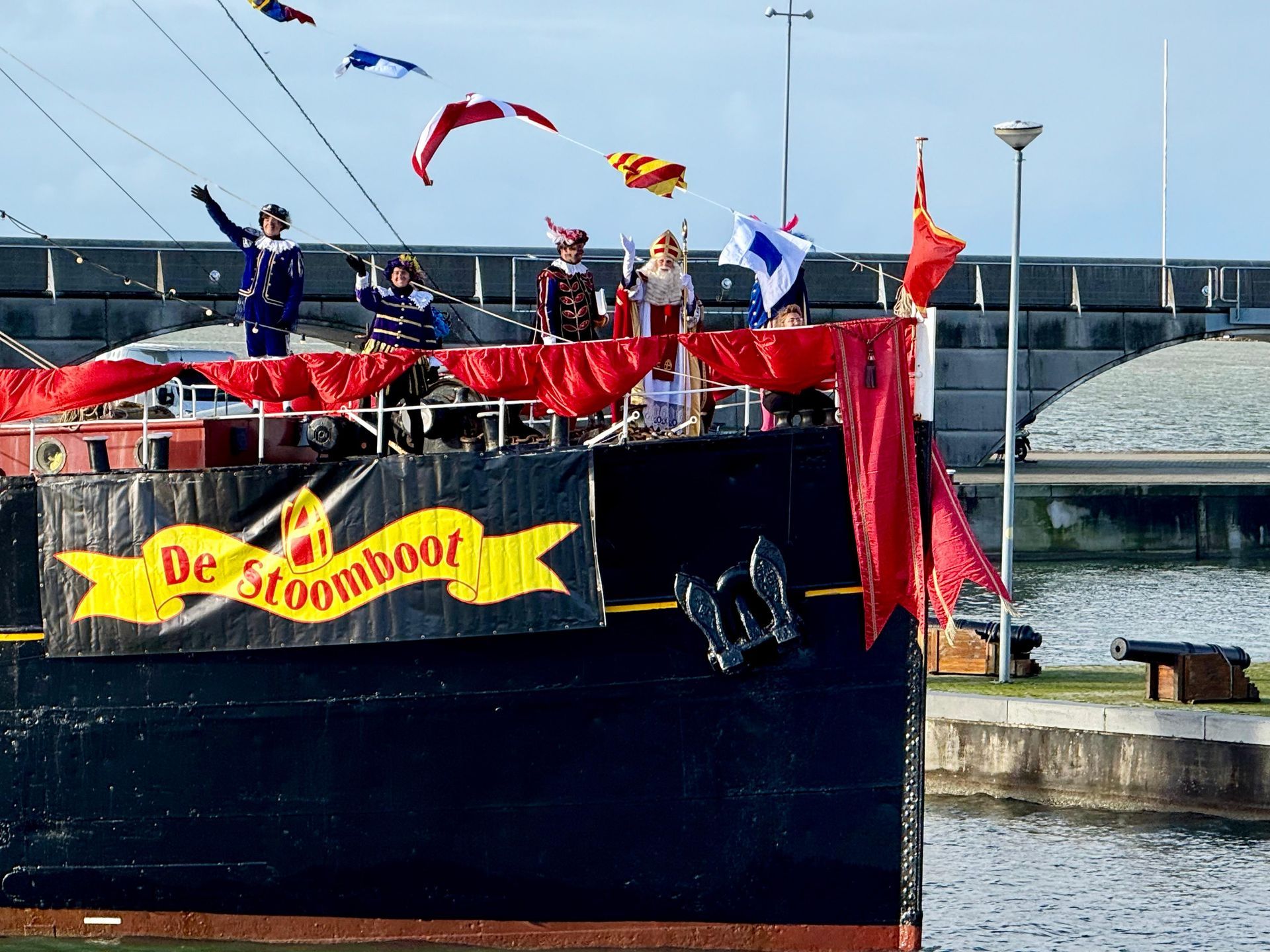 Een zwarte boot genaamd De Stoomboot, versierd met rode draperieën en feestelijke vlaggen, met verklede figuren die naar de menigte zwaaien.