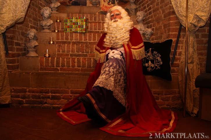 Sinterklaas, gekleed in een traditioneel rood gewaad met gouden versieringen, zit in een schemerig verlichte kamer met bakstenen muren en decoratieve beelden.