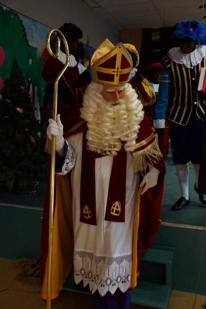 Een persoon in traditionele Sinterklaaskleding, met mijter en gewaad, die een staf vasthoudt, met metgezellen op de achtergrond.