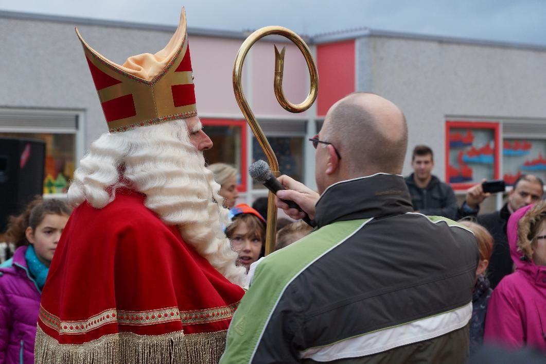 Een persoon, verkleed als Sinterklaas in rode gewaden en met een mijter op zijn hoofd, spreekt in een microfoon die door een interviewer buiten wordt vastgehouden.
