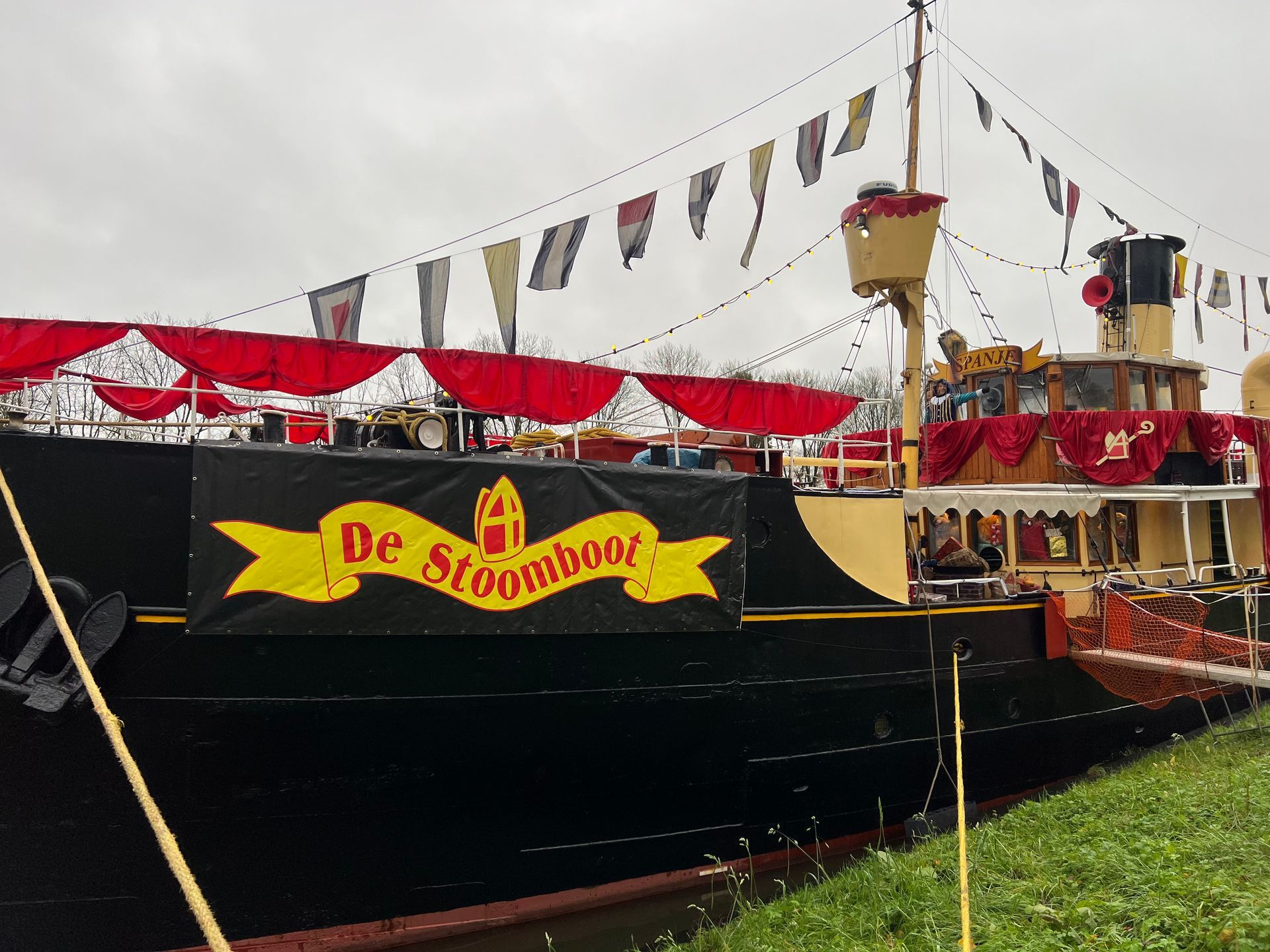 Een zwarte stoomboot versierd met rode slingers en vlaggen, met een geel spandoek waarop 