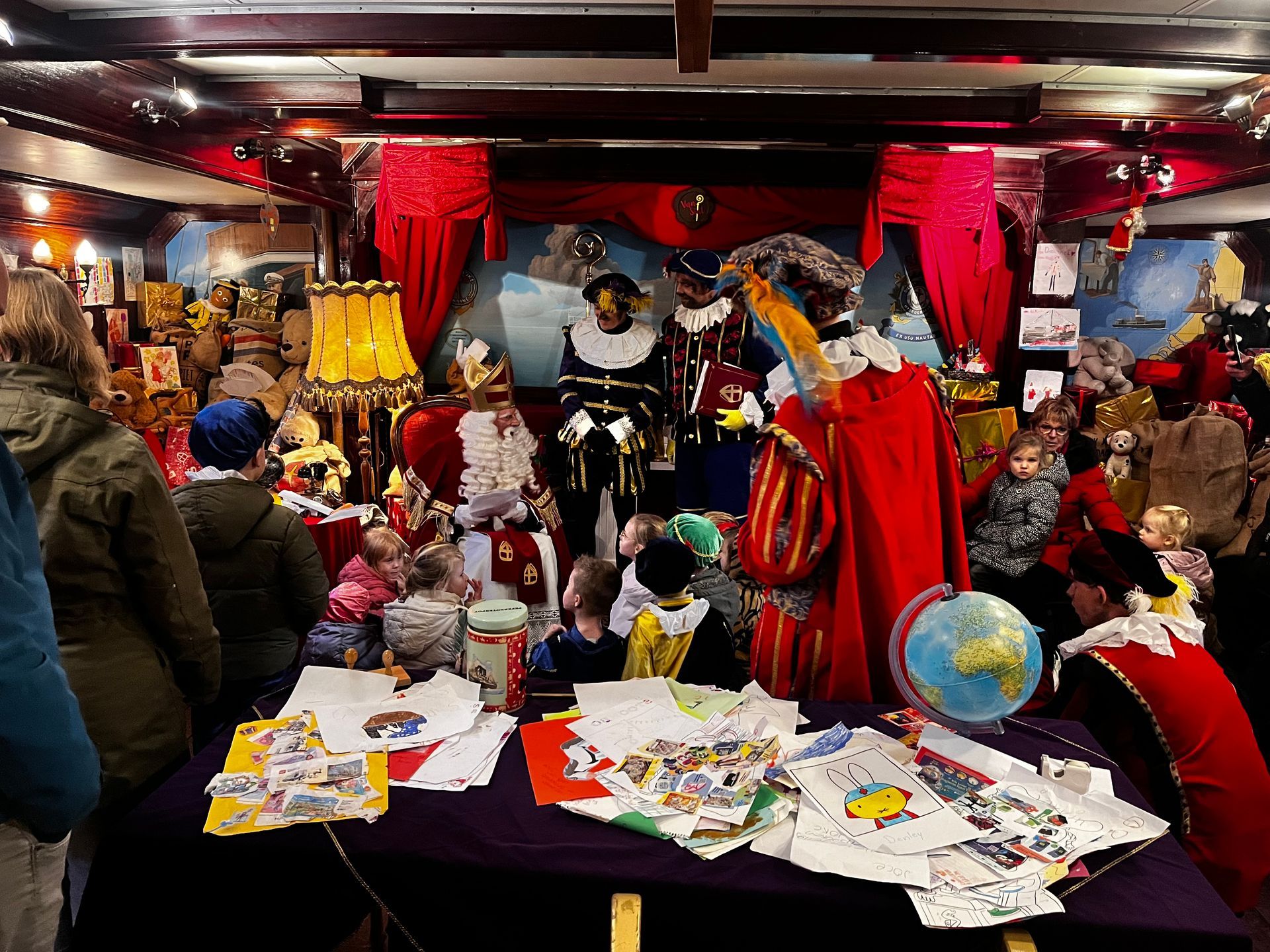 Sinterklaas en Zwarte Pieten begroeten kinderen in een versierde kamer met tafels vol tekeningen en een wereldbol.