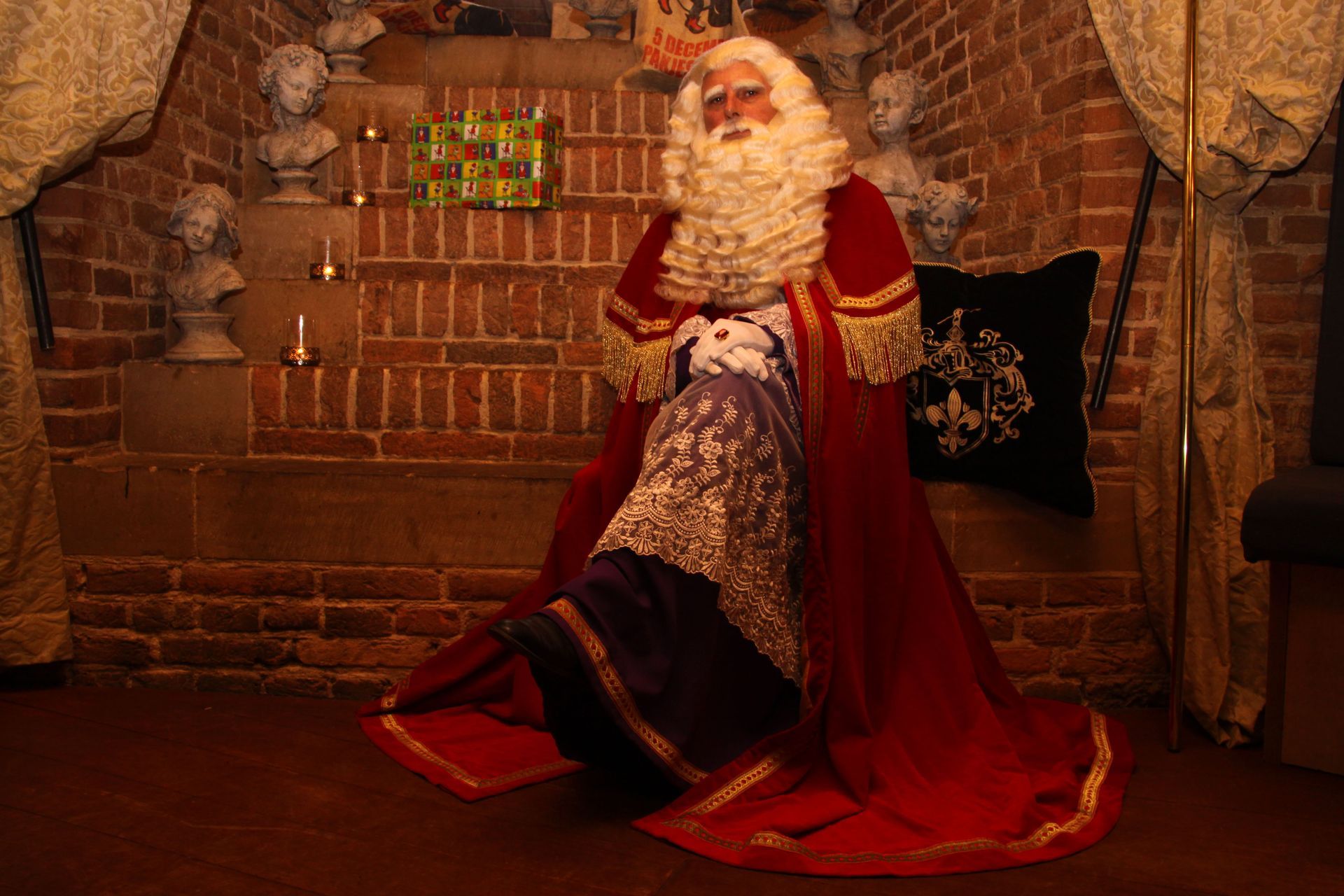 De kerstman zit op een trede in een kamer met bakstenen muren, gekleed in een rode fluwelen cape en met een klein knuffeldier in zijn handen.