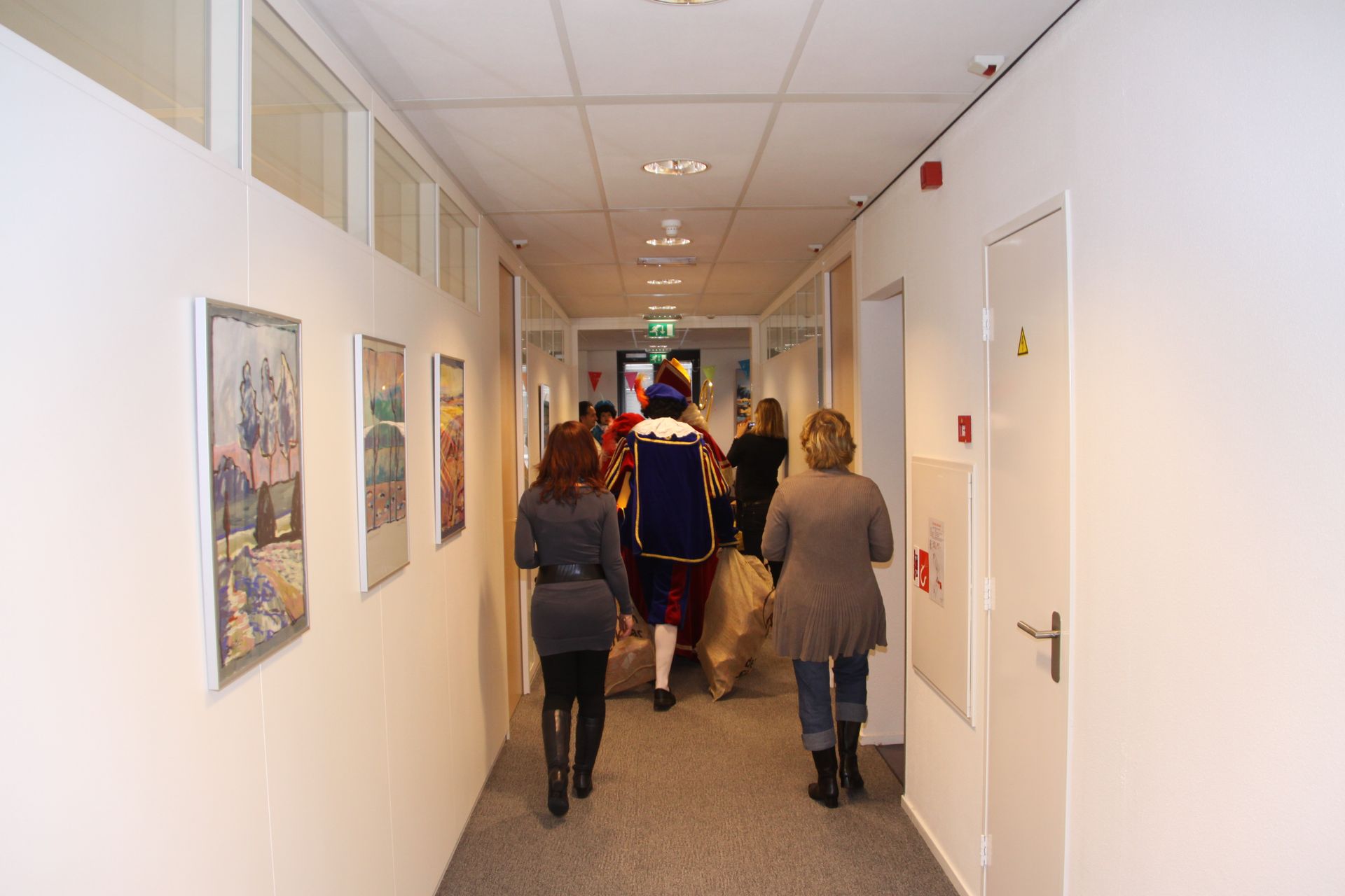 Mensen lopen door een gang in de richting van een figuur gekleed in een kleurrijk historisch kostuum, die een grote papieren tas draagt.