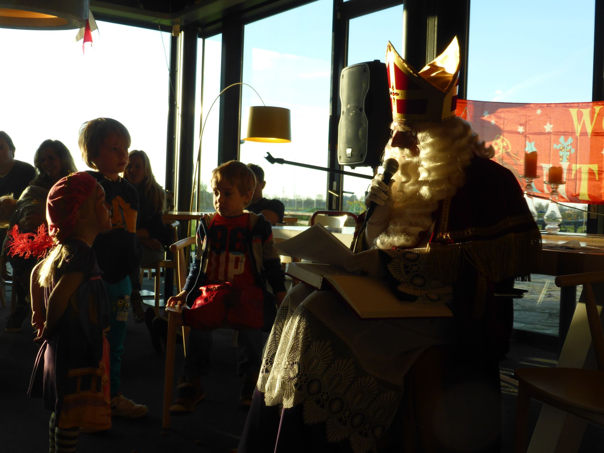 Sinterklaas zit binnen voor te lezen aan kinderen in een verlichte kamer, met versieringen zichtbaar op de achtergrond.