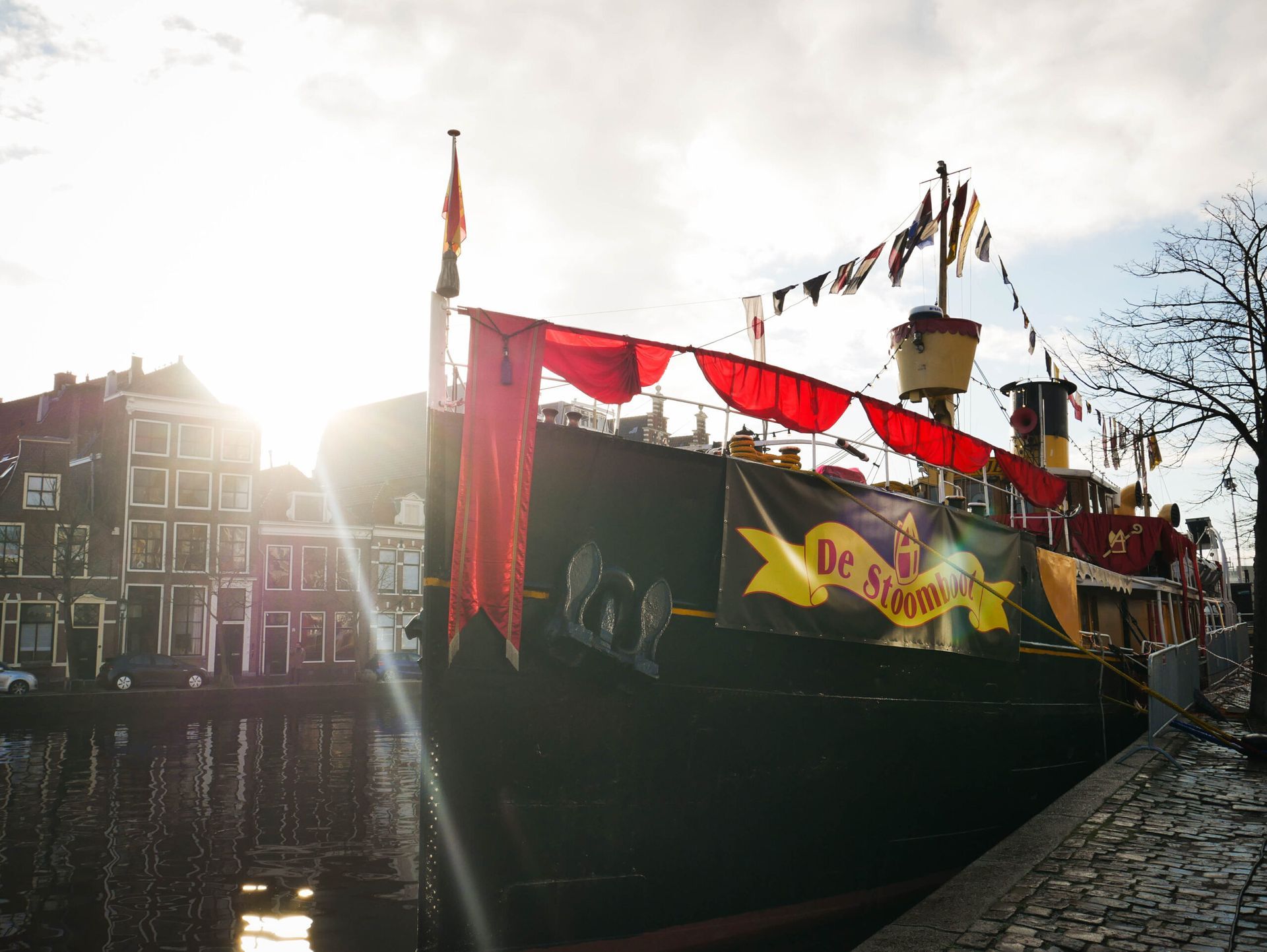 Een zwarte boot, versierd met rode stof en vlaggen, ligt bij zonsondergang op een kanaal, met gebouwen op de achtergrond.