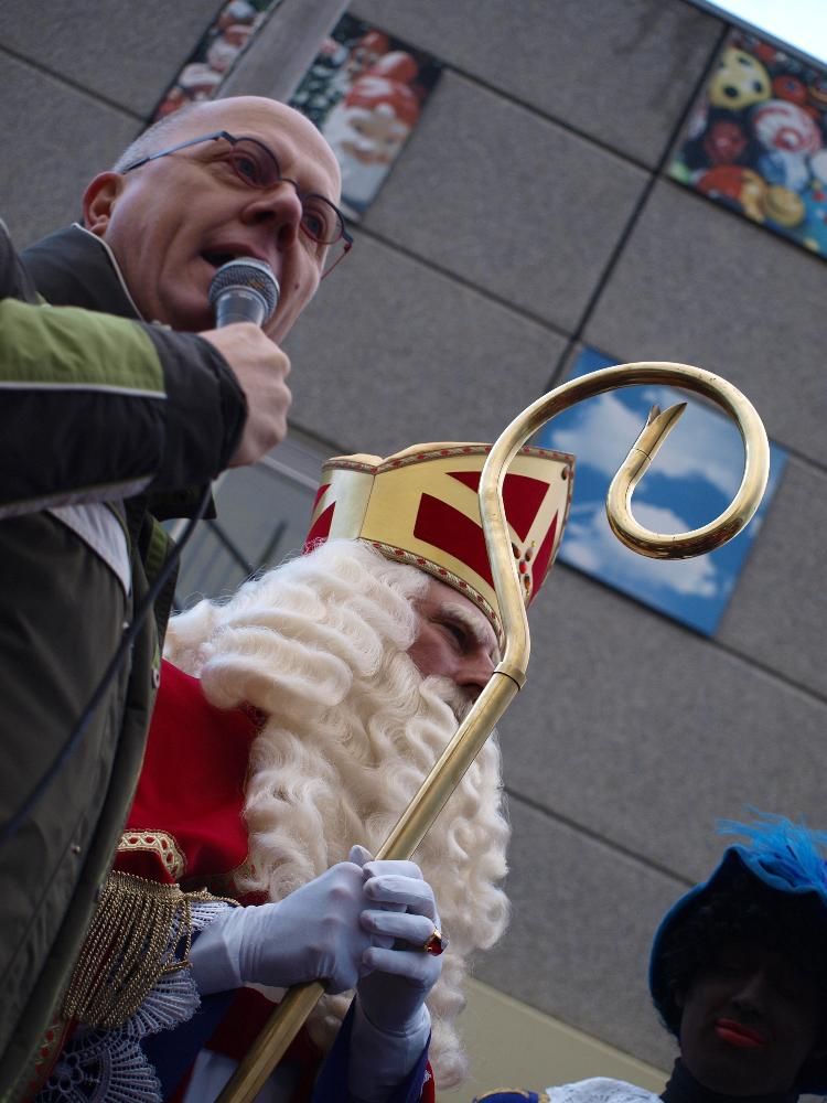 Naast Sinterklaas, die een gouden staf vasthoudt, staat een spreker met een microfoon, en een helper met een blauwe muts.