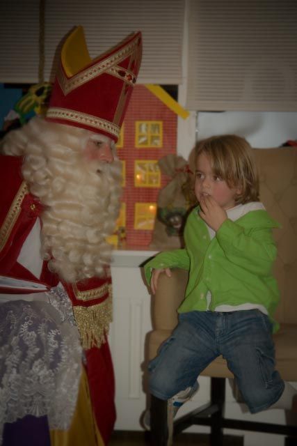 Een persoon in een rood-gouden Sinterklaaskostuum staat naast een zittend kind in een felgroene trui.