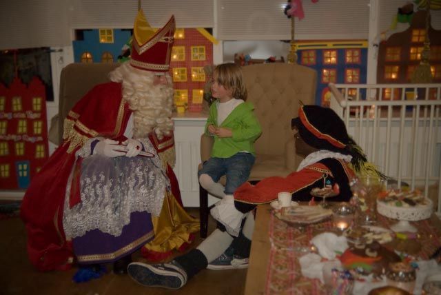 Een kind zit tussen Sinterklaas en Zwarte Piet in een kamer die versierd is met miniatuur speelgoedhuisjes.