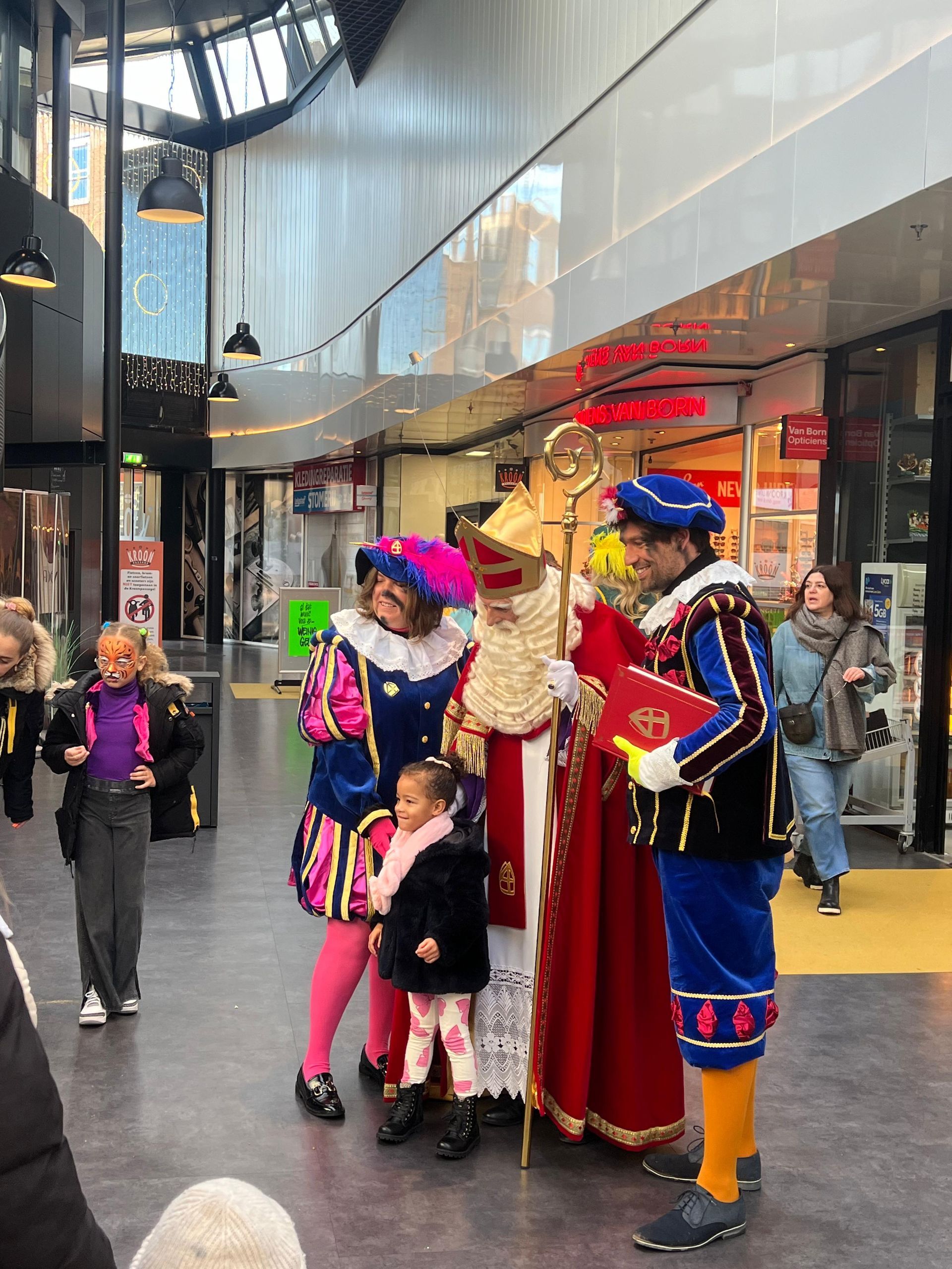 Sinterklaas en twee helpers in traditionele klederdracht staan ​​in een winkelcentrum en praten met een jonge bezoeker.