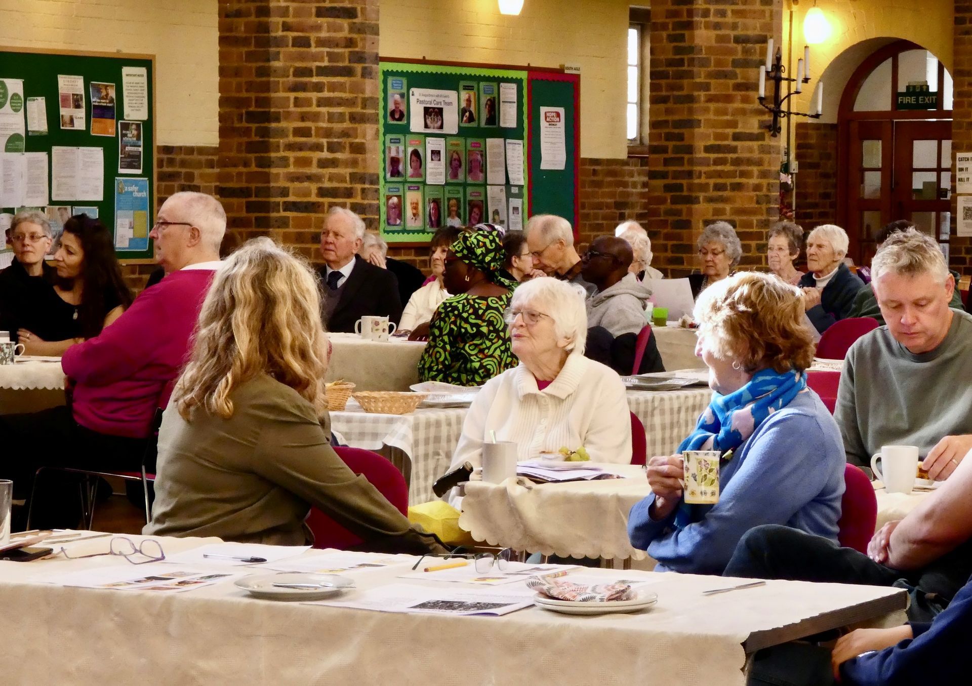 St Augustine Cafe Church. People seated at tables in a brick-walled community hall, chatting during an indoor gathering