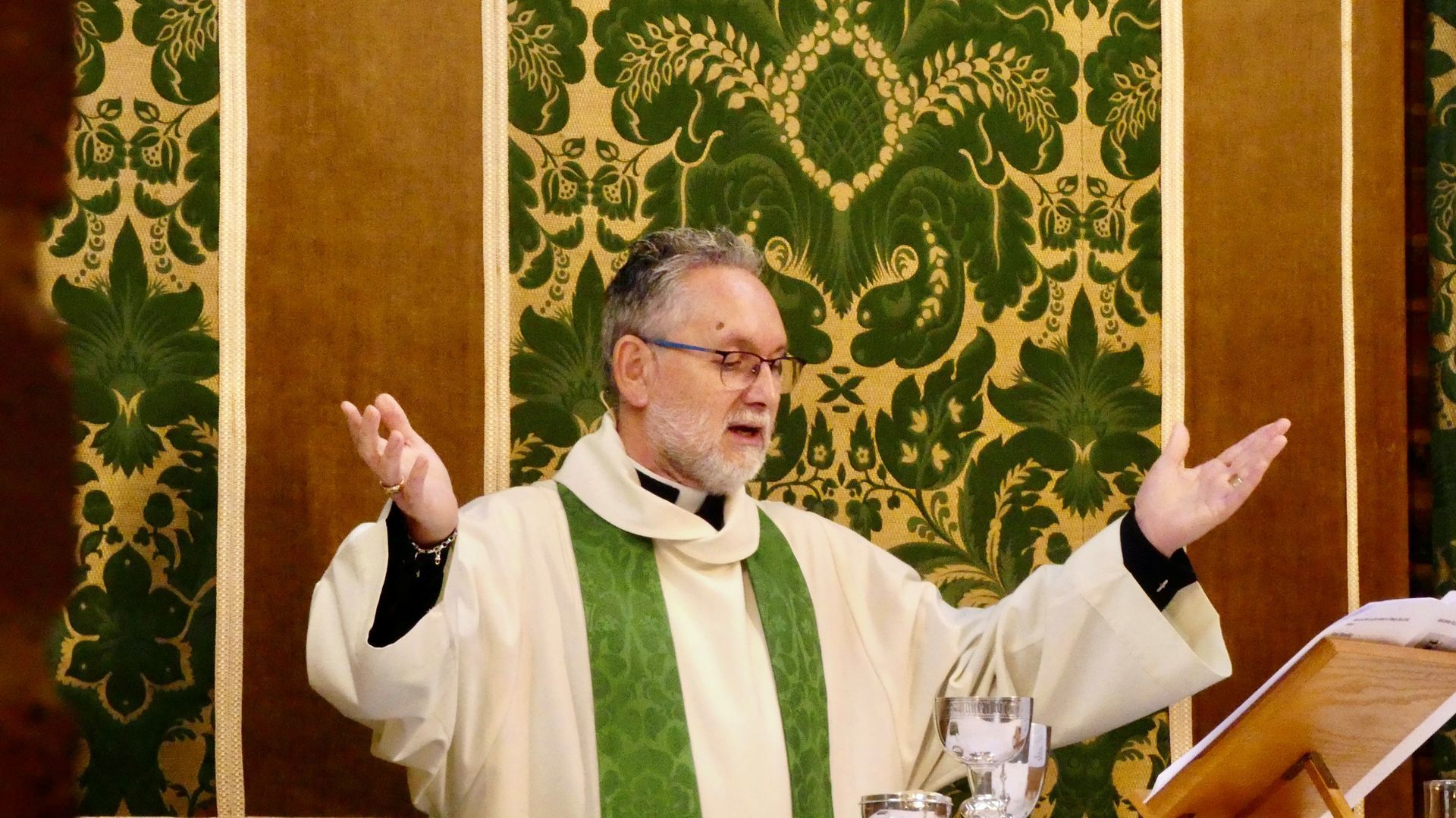 Priest in green vestments with raised hands preaching at an altar in a church.