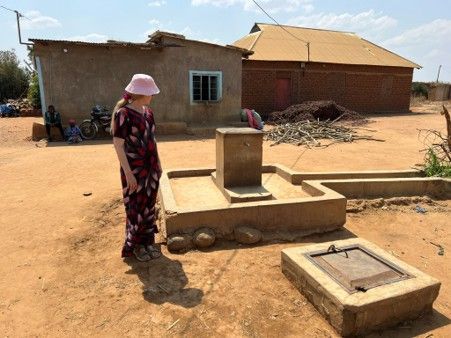 Woman standing beside a concrete water pump in a sandy rural yard, with simple houses in the background.