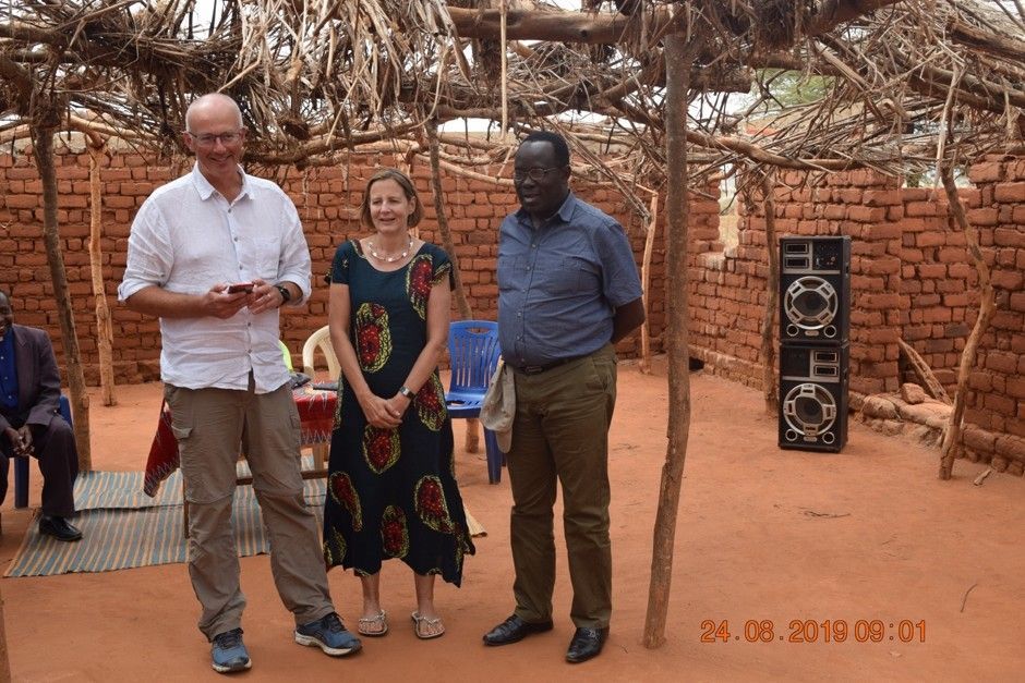 Three people standing in a rustic outdoor courtyard with a brick wall and speaker stack in the background.