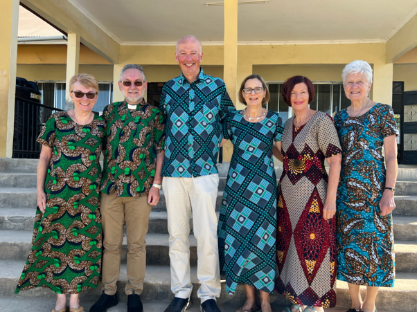 Six people stand together outdoors in colorful patterned outfits, smiling in front of a building.