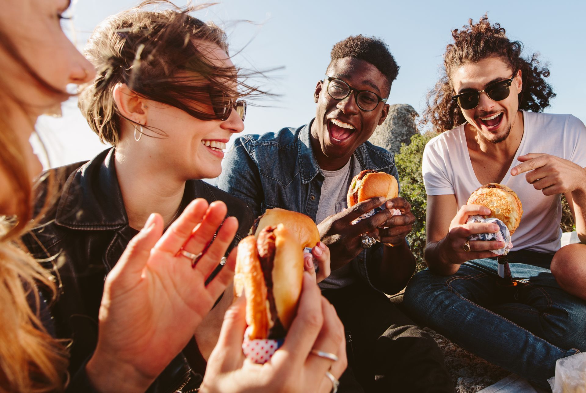 Four people laughing and eating burgers outdoors.