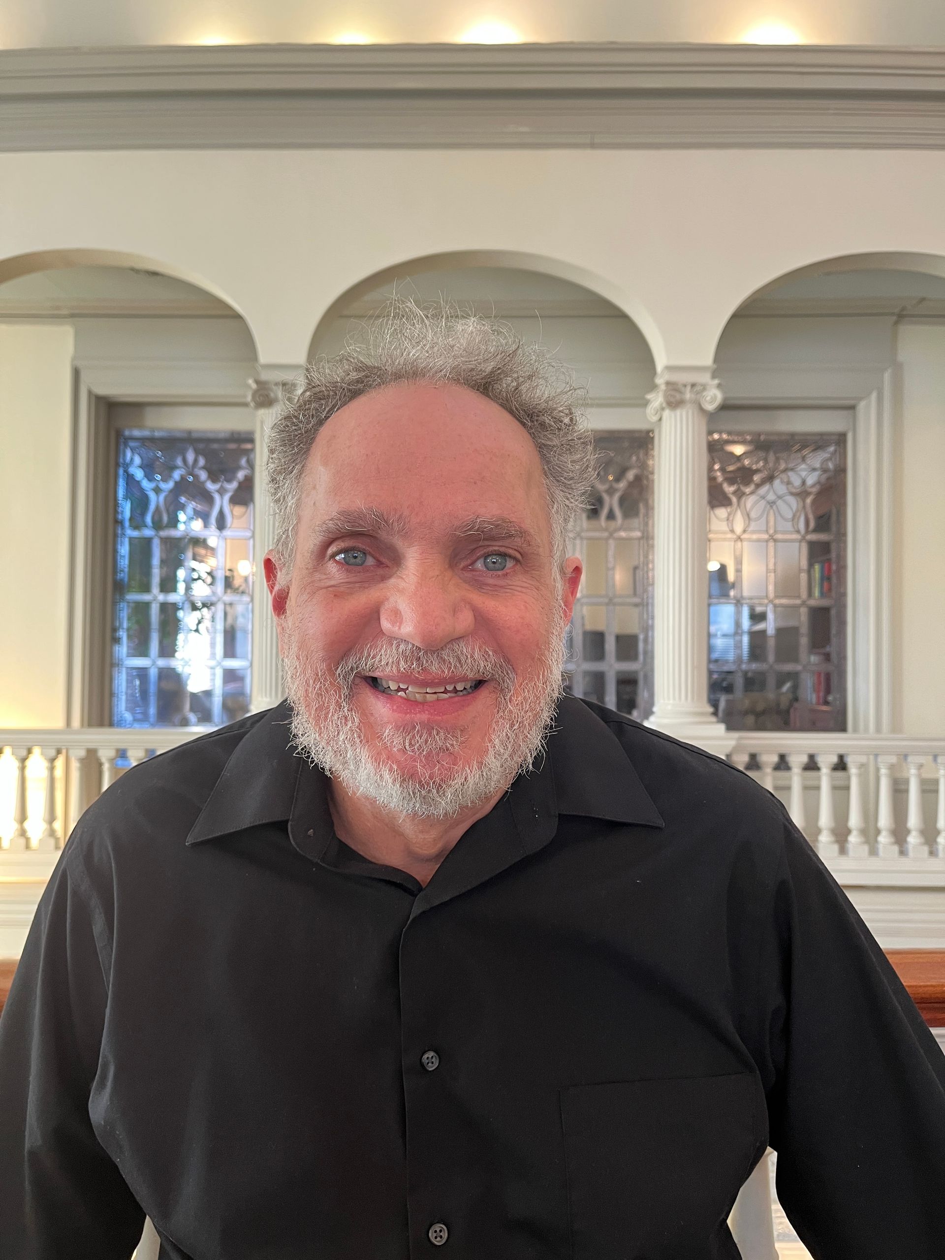 Man with beard smiles, wearing black shirt, in a building with windows and white columns.
