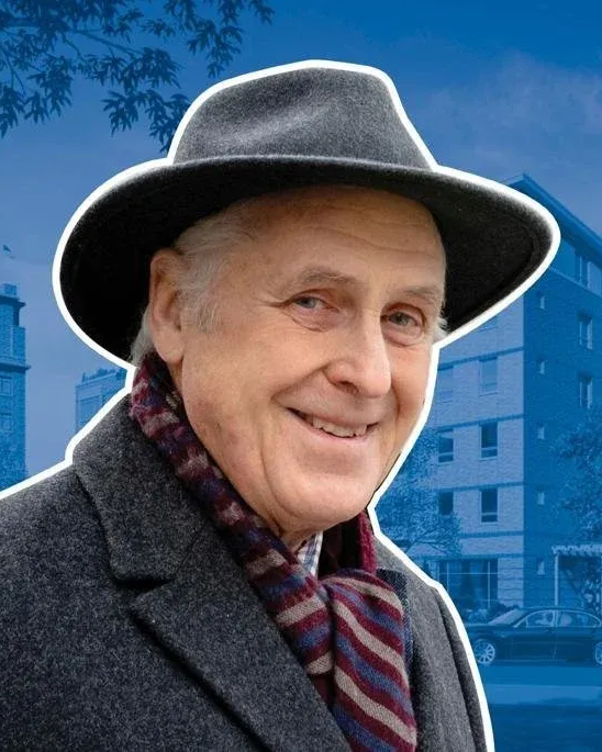 Man wearing a fedora and scarf, smiling; building backdrop.