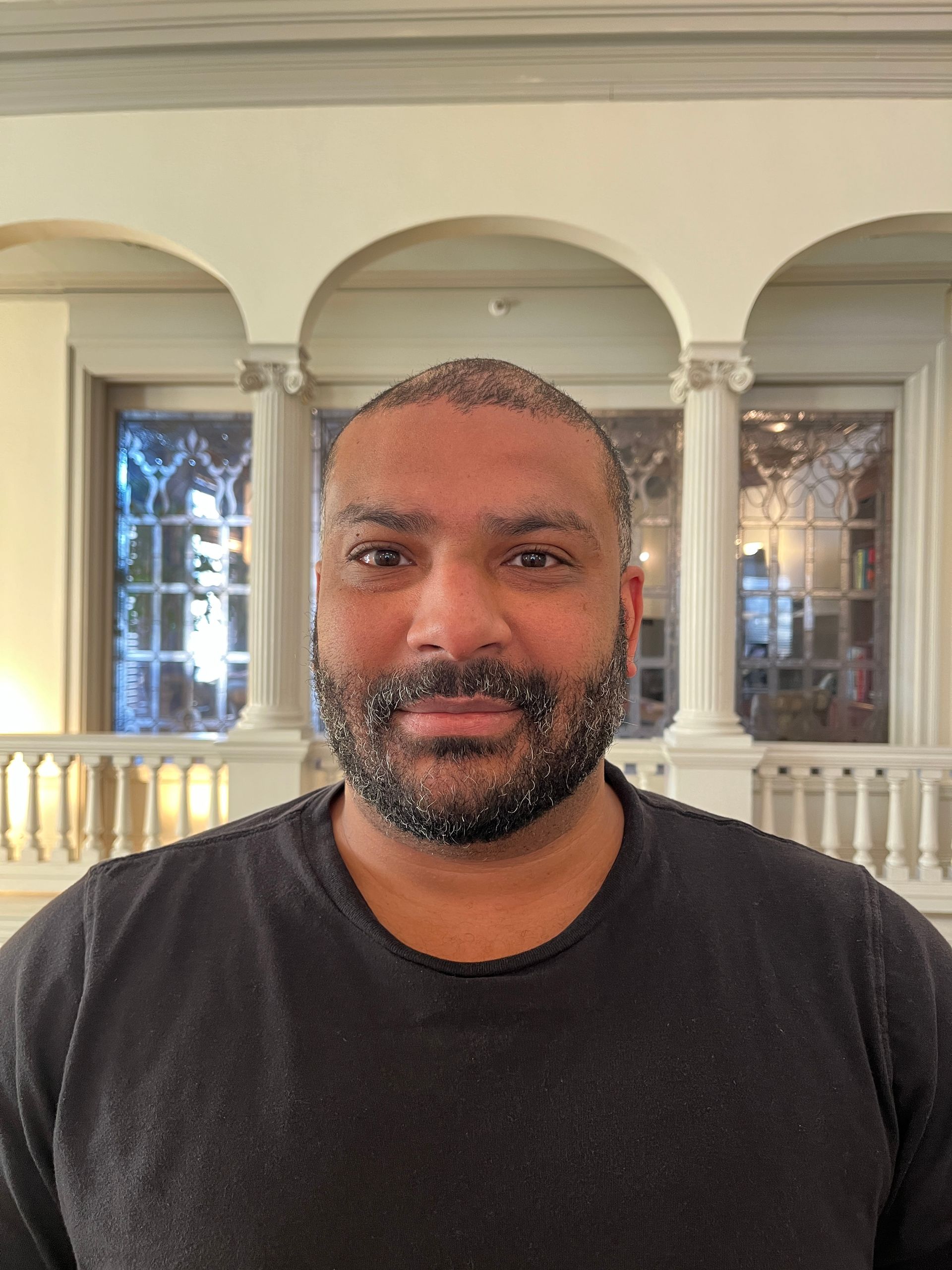 Man with a beard in a black shirt, in a building with columns and arches.