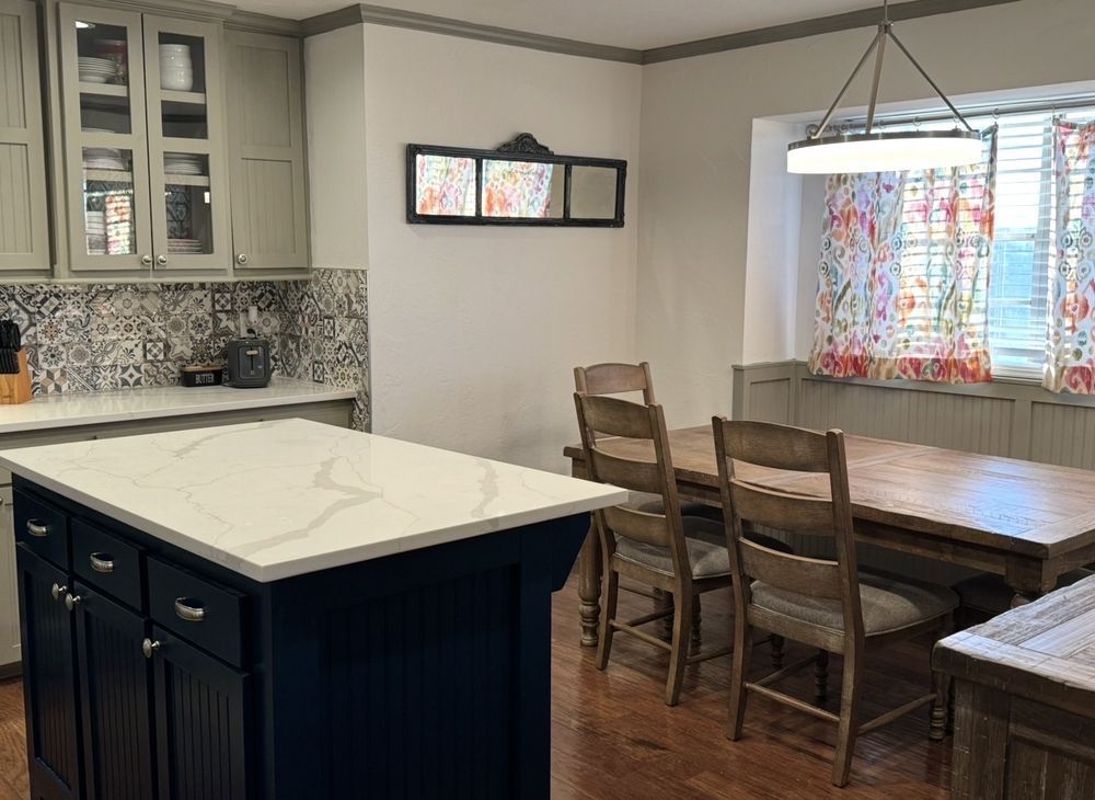 Bright kitchen with navy island, white counters, and a wooden dining table by a window with patterned curtains