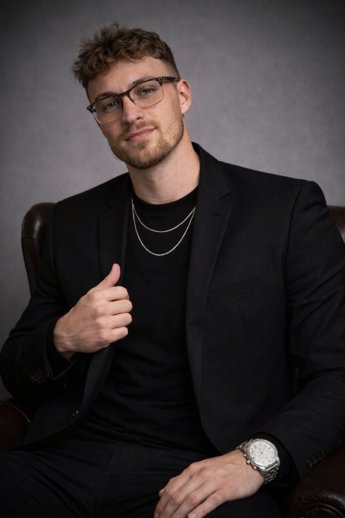 Portrait of a man in a black suit and glasses, seated against a gray studio backdrop, wearing layered necklaces.