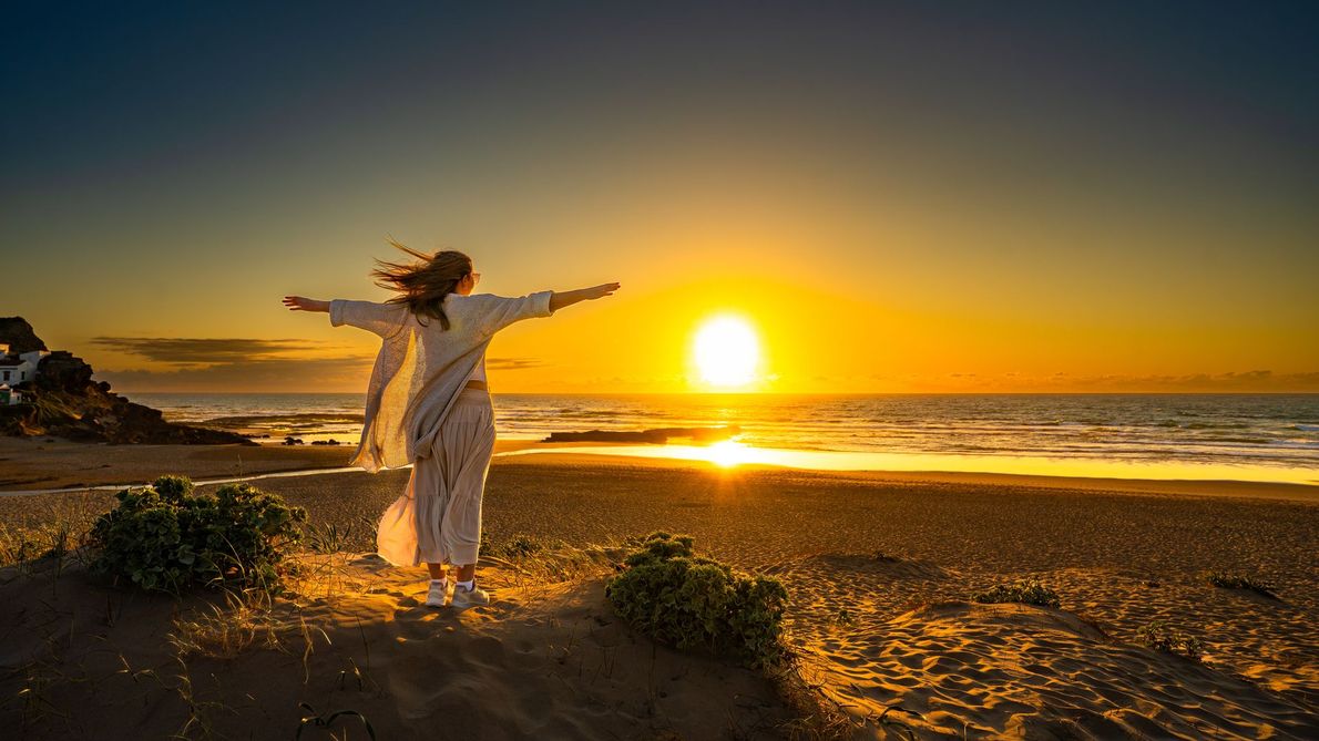 Person in a white dress stands on a beach at sunset with arms outstretched.
