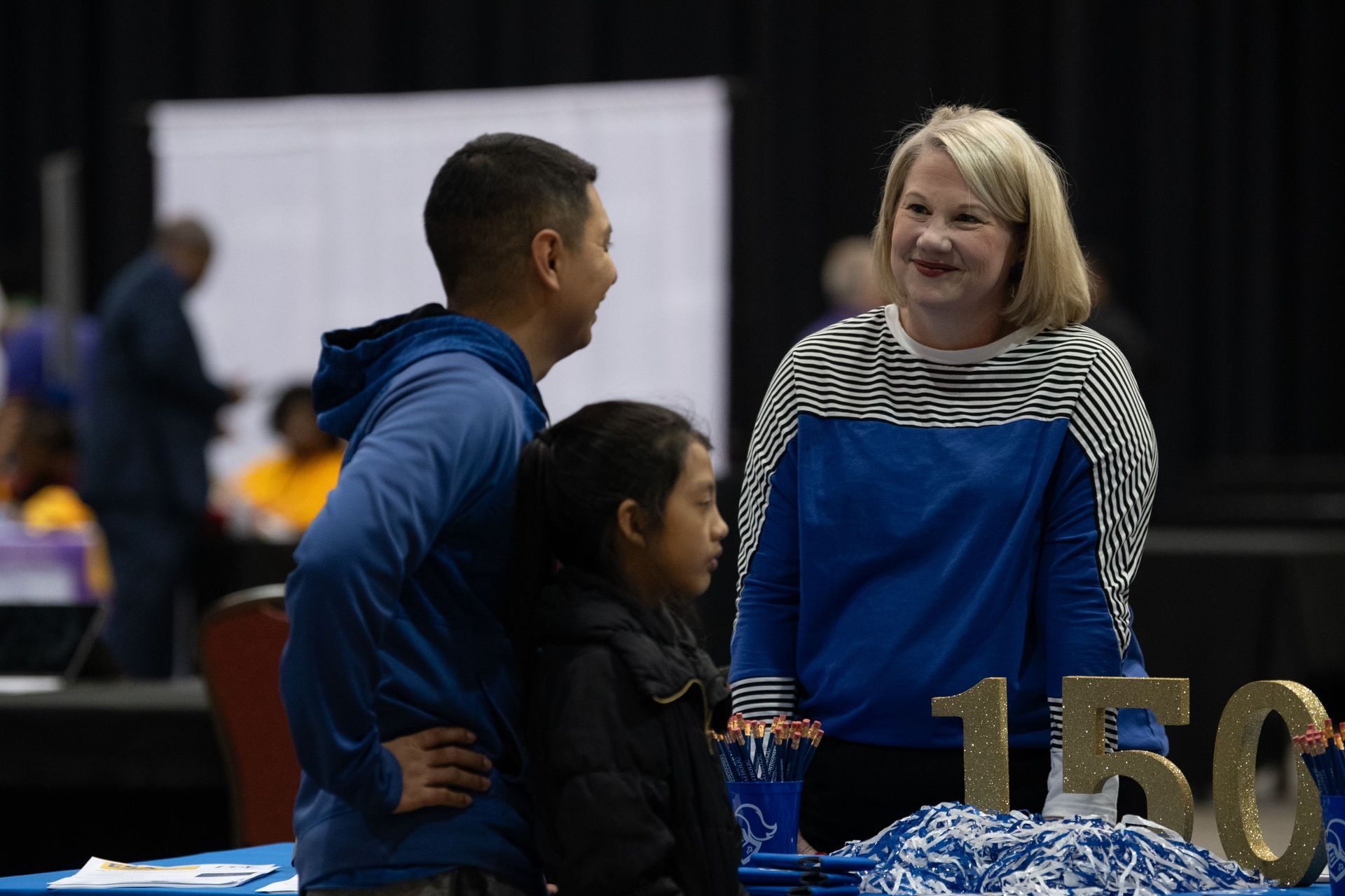 Three people talking at an indoor event table with a gold “150” display in the foreground.