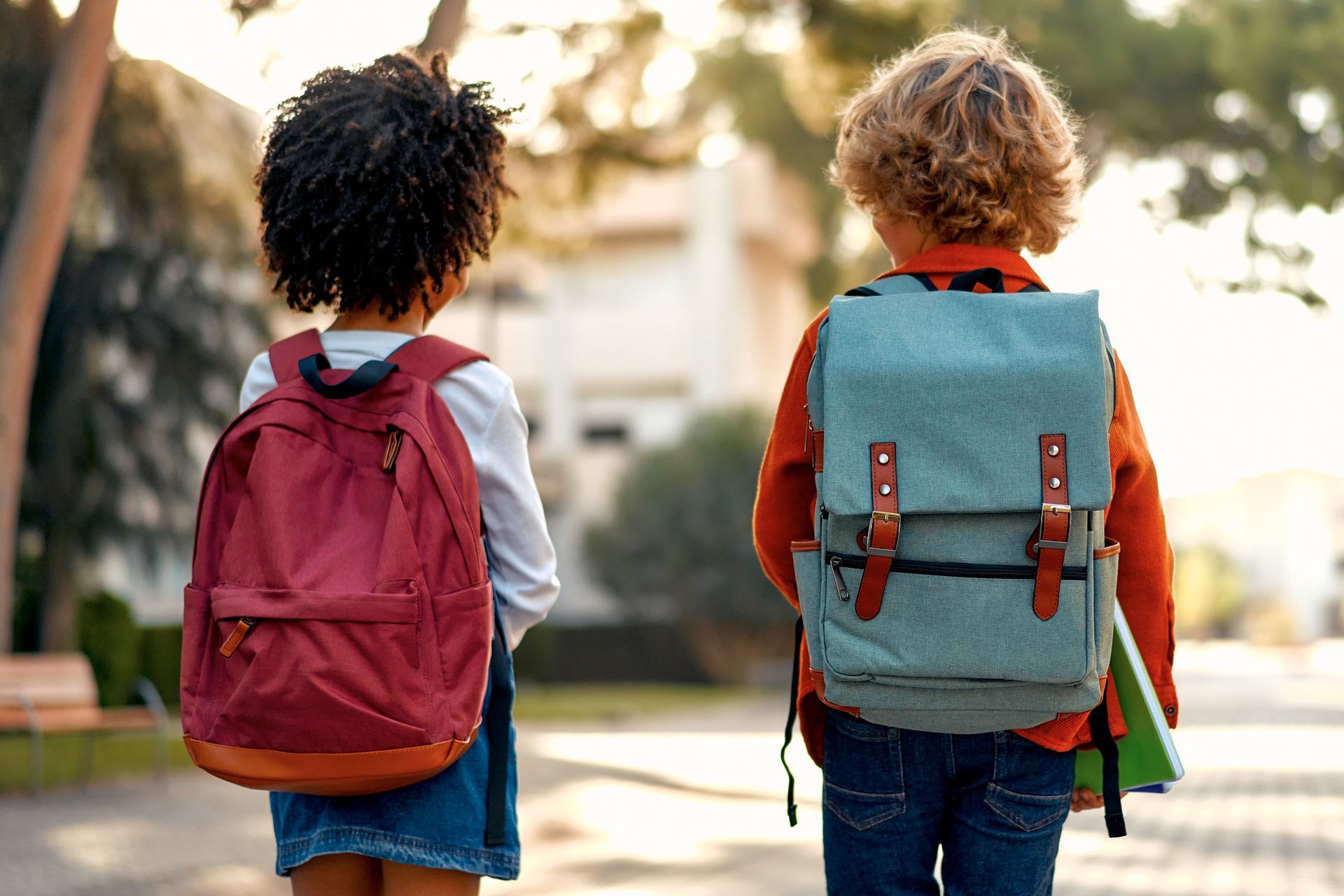 Two children with backpacks walk away from the camera toward a school building on a sunny day.