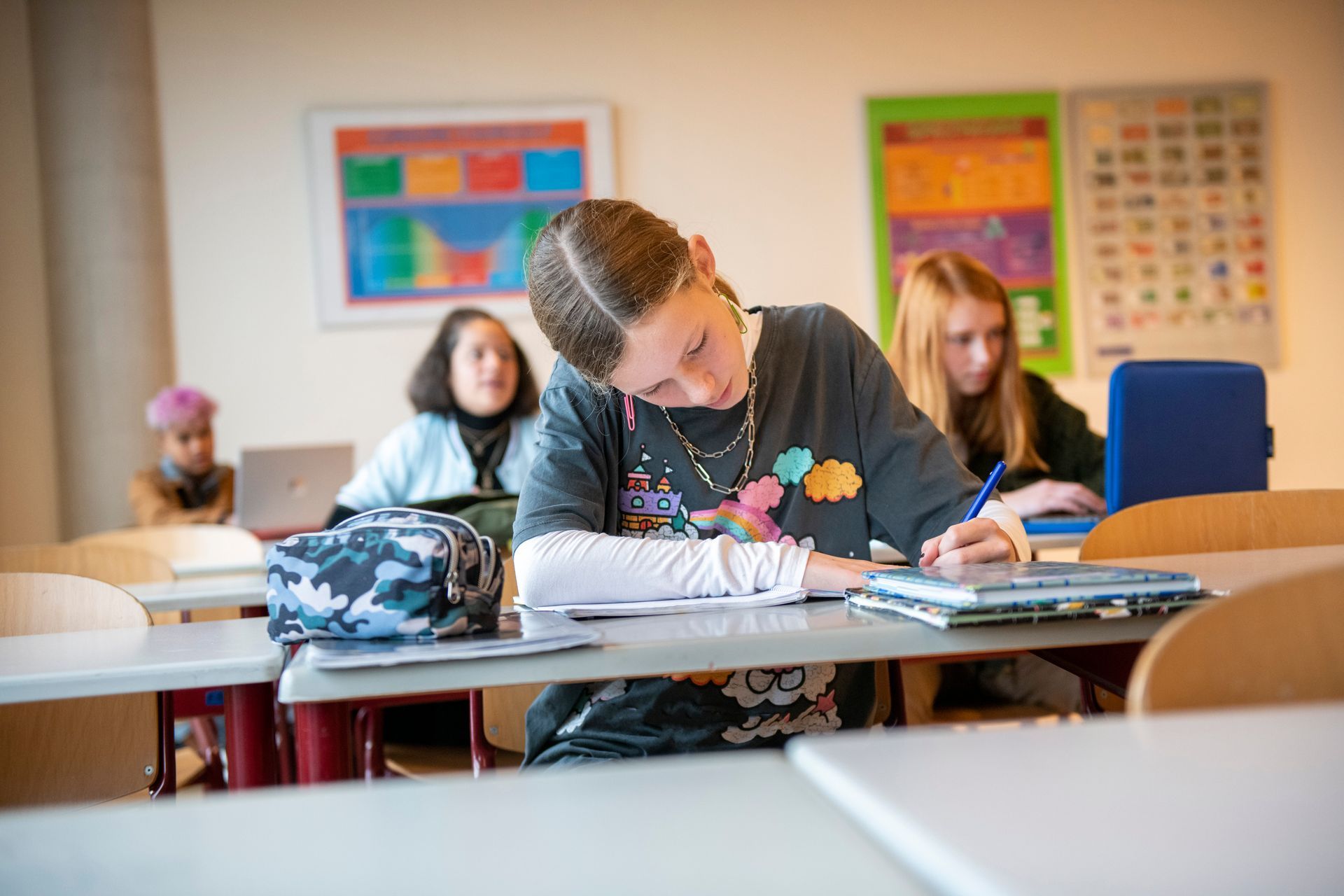 Student writing at a classroom desk while classmates sit behind them, with colorful posters on the wall.
