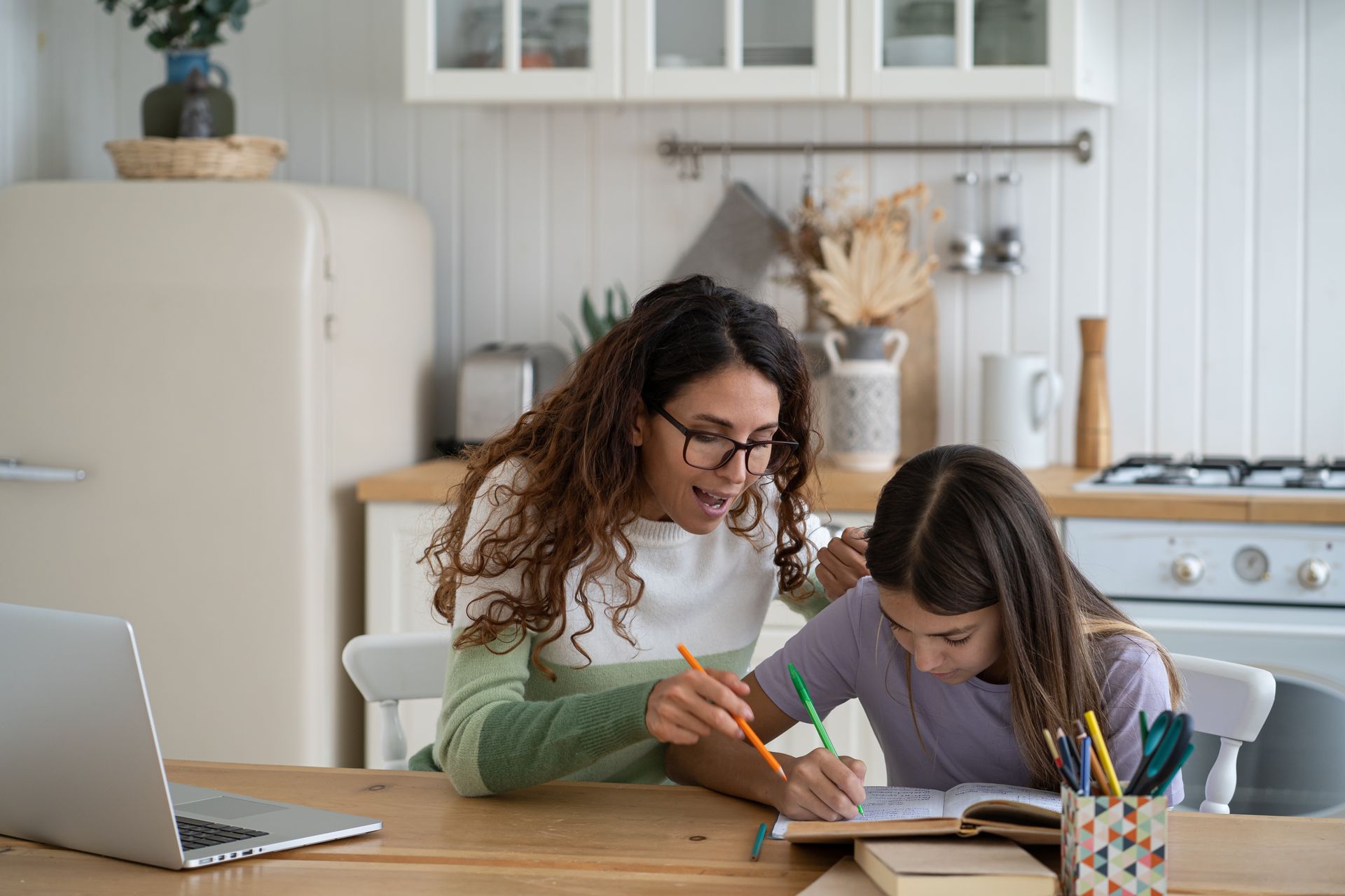 A mom helping child with school work at a kitchen table, with a laptop and pencil cup nearby