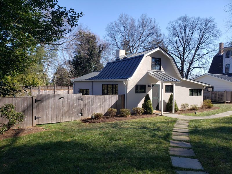 Small white house with dark roof, fenced yard, and stone path under a clear blue sky
