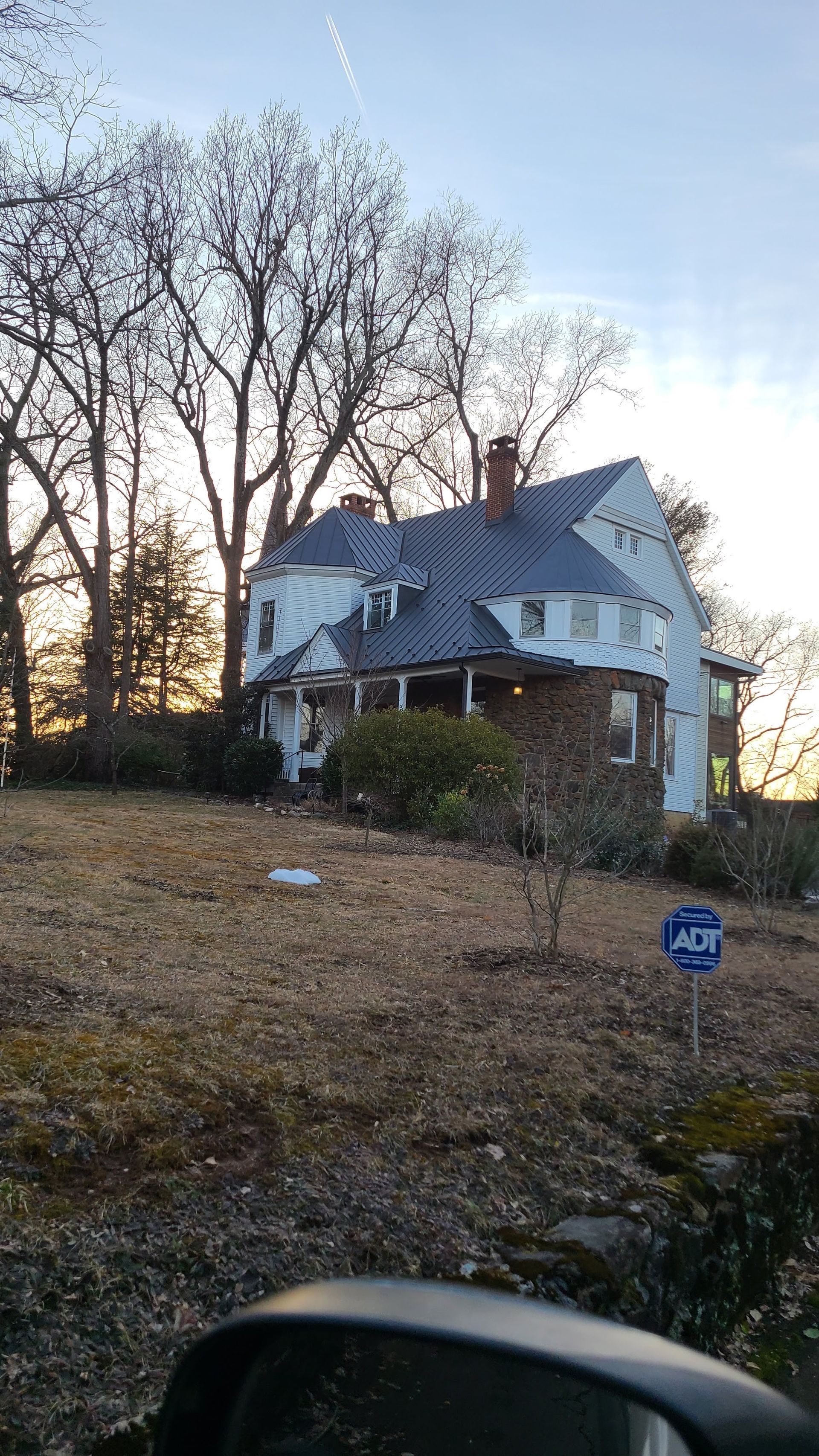 Old brick house with a steep gray roof on a grassy lot, with leafless trees at sunset.