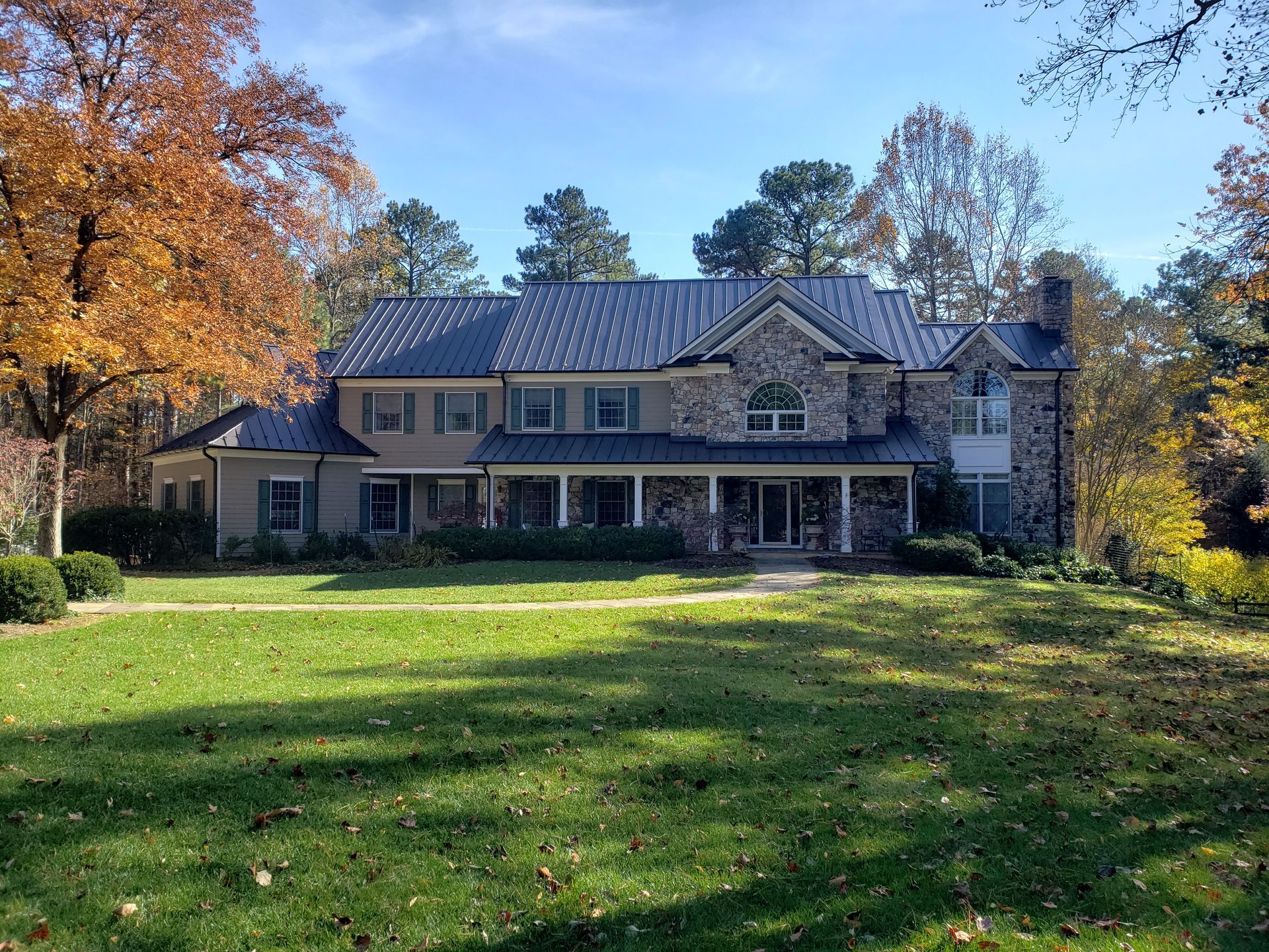 Large stone house with a covered porch, surrounded by autumn trees and a green lawn under a blue sky