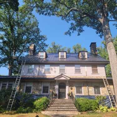 Three-story house with a front staircase and ladders on both sides, framed by tall trees under a blue sky