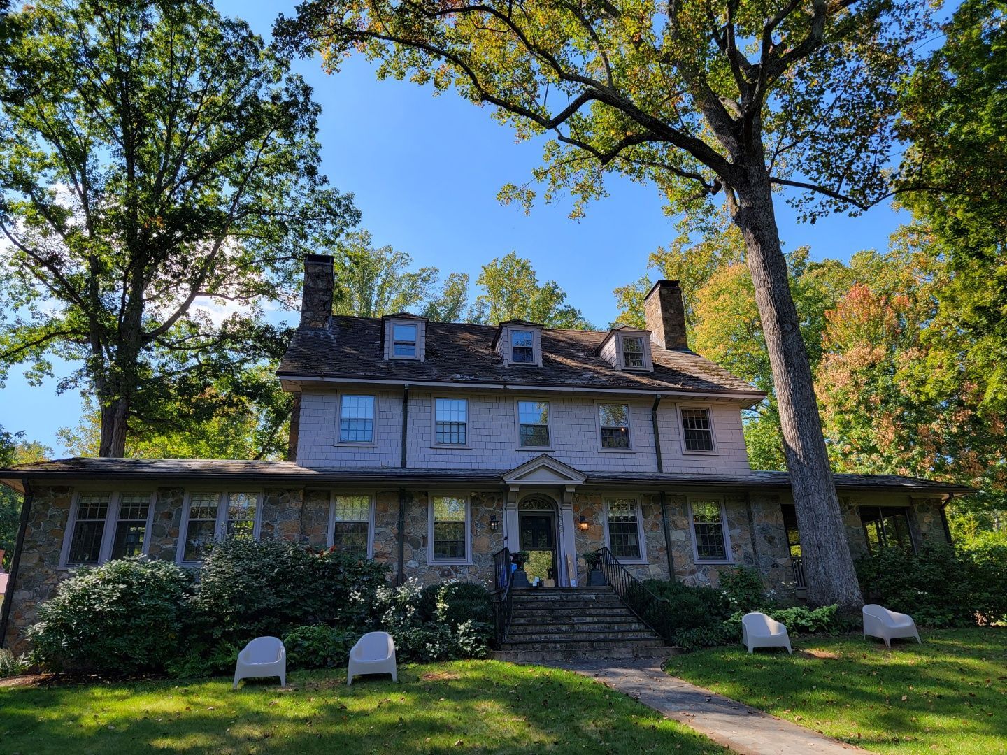 Old stone house with a porch, framed by tall trees and a green lawn under a bright blue sky