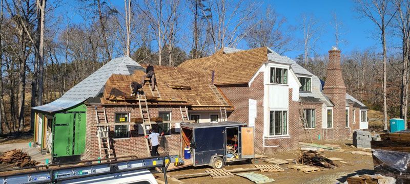 House under construction with workers, scaffolding, and a partially finished roof on a wooded lot.