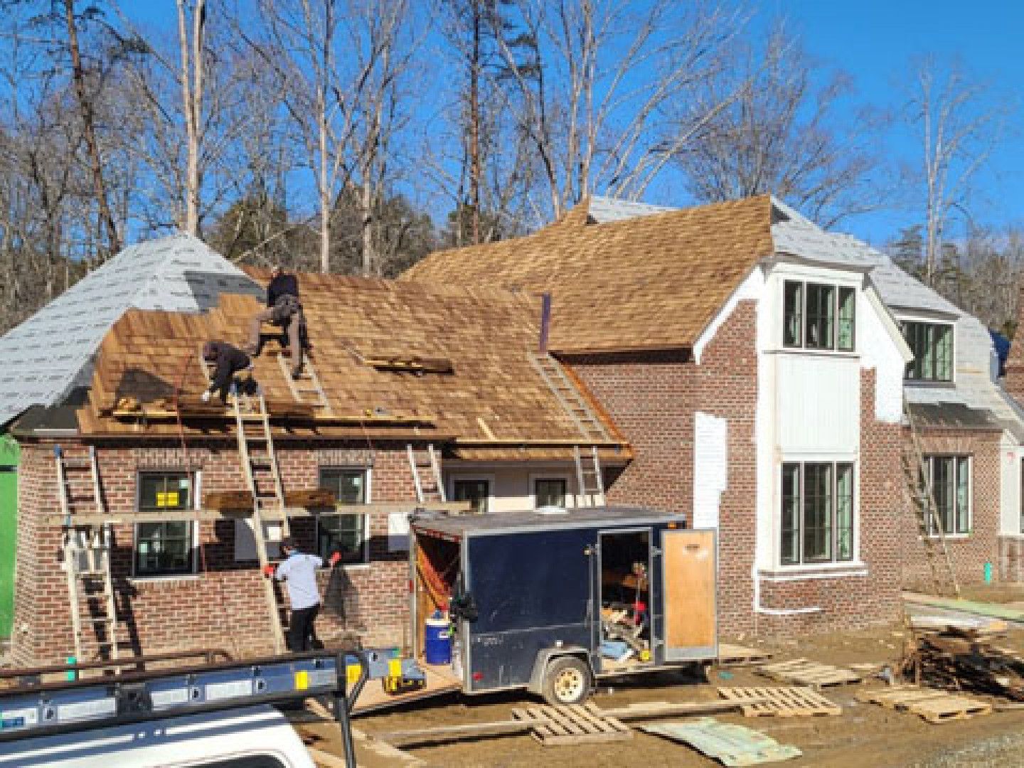 Workers replacing a roof on a brick house with a white dormer and a trailer in the driveway