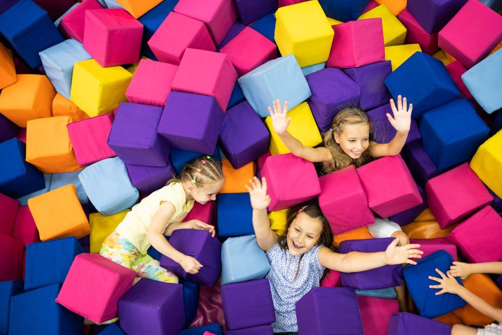 Three children laughing while playing among a pile of colorful, large foam blocks in a play area.
