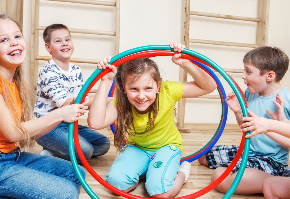 Children sit on a wooden floor in a gym, laughing as they hold up and pose behind large, colorful circular hoops.