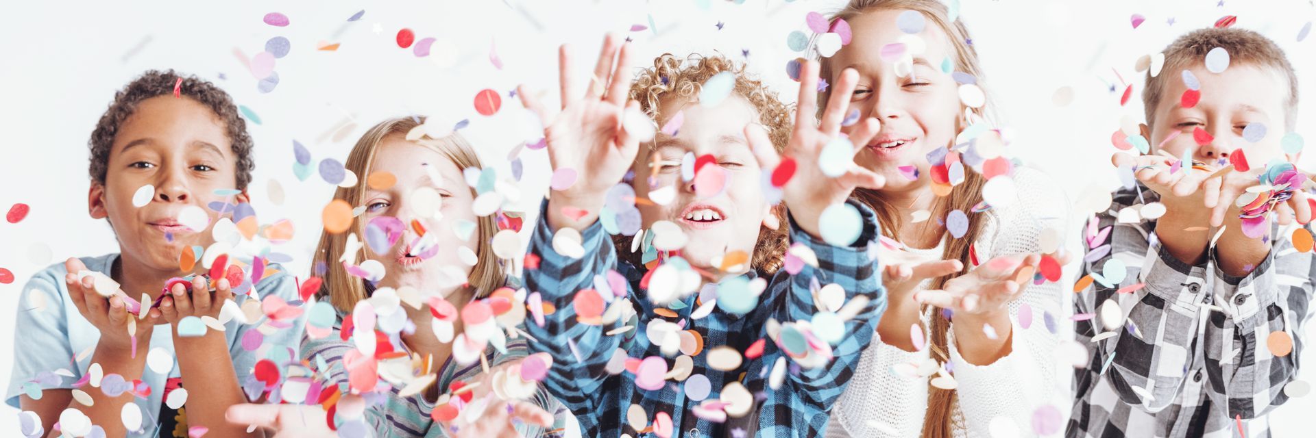 Five children stand in a row against a white background, happily blowing colorful confetti from their cupped hands.