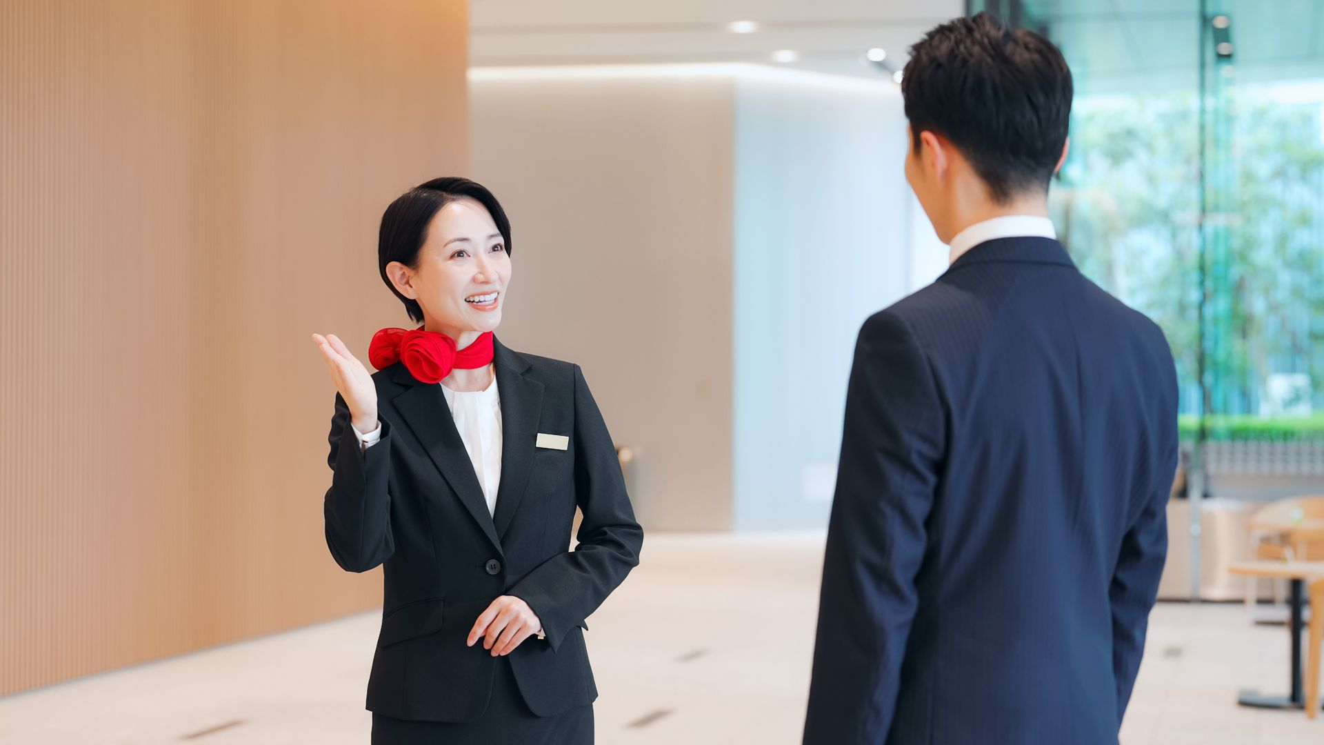 Hotel staff in black suit with red scarf greeting a guest in a bright lobby.