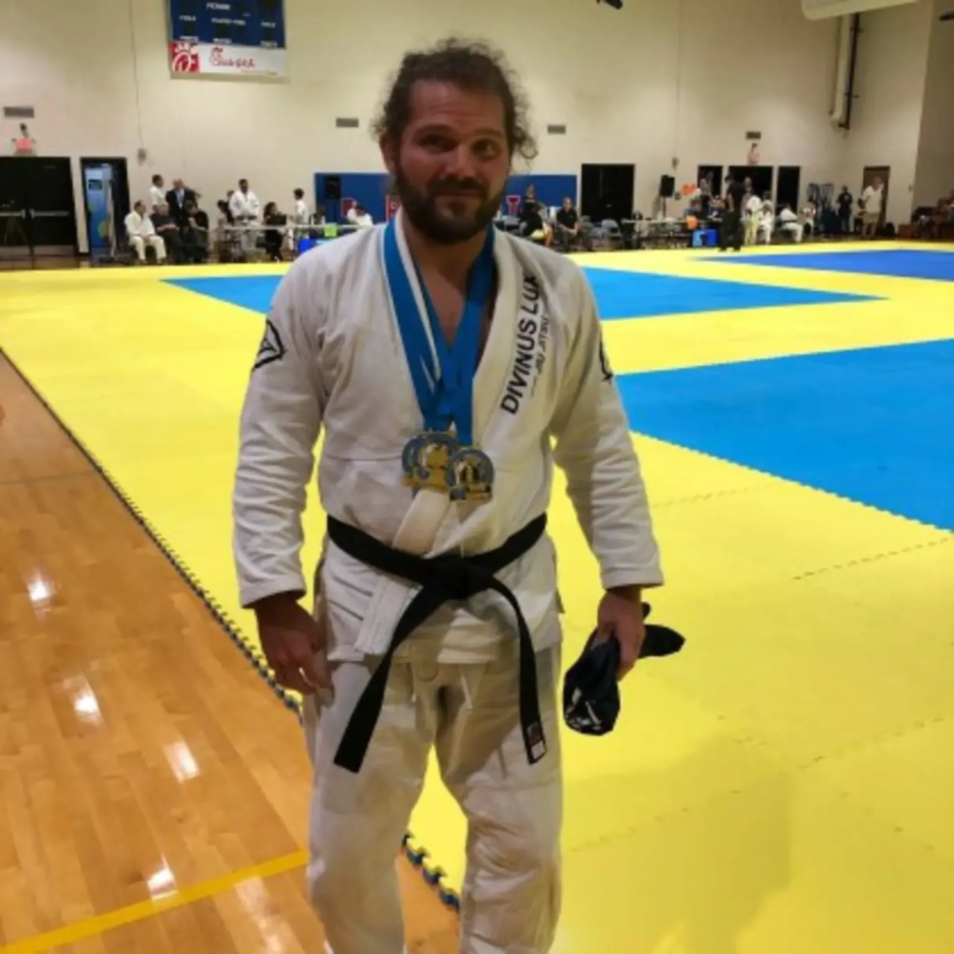 Jiu-jitsu competitor in white gi with medals stands on a yellow-and-blue mat after a match.