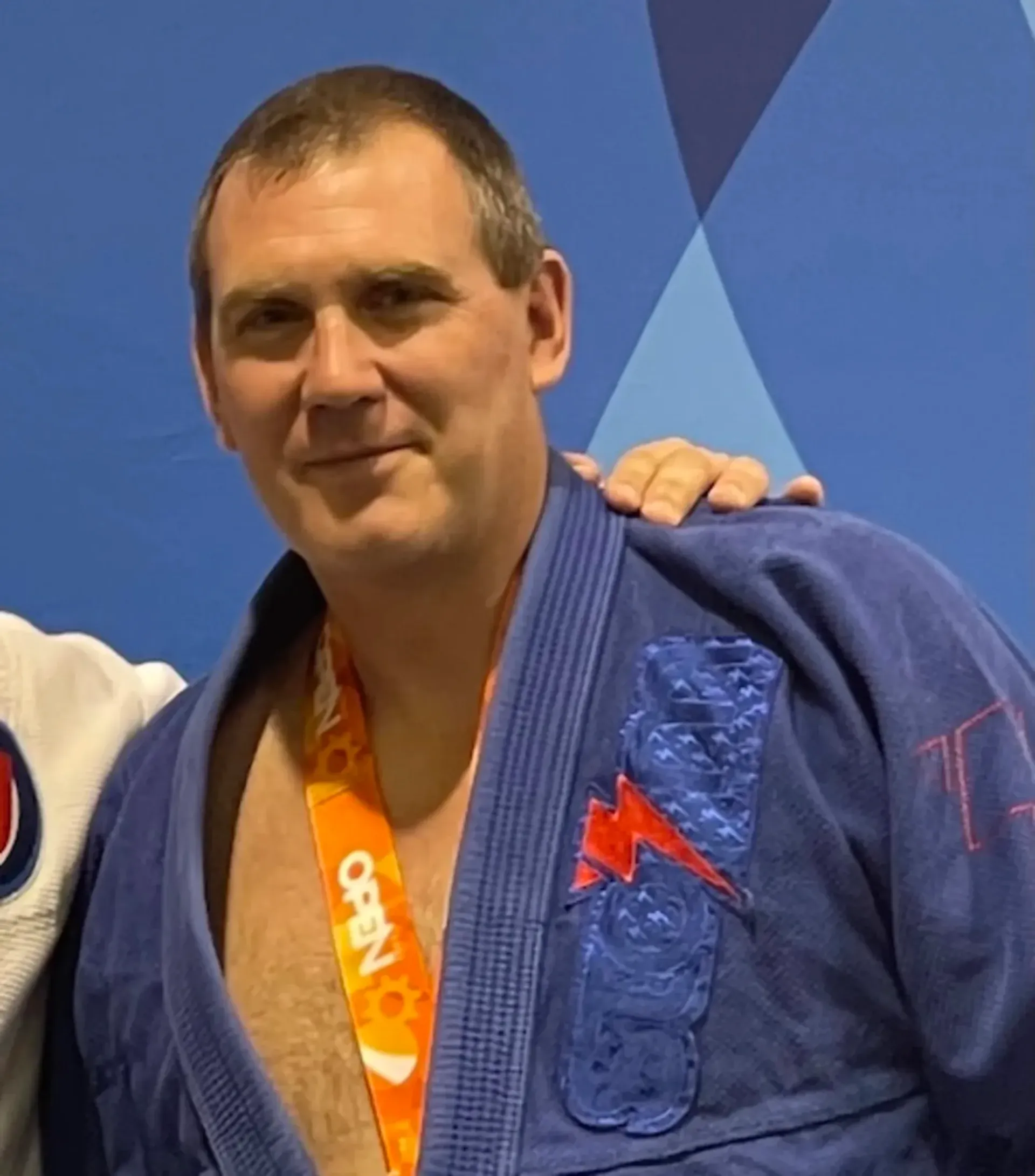 Man in a blue gi with orange lanyard smiles against a blue patterned backdrop