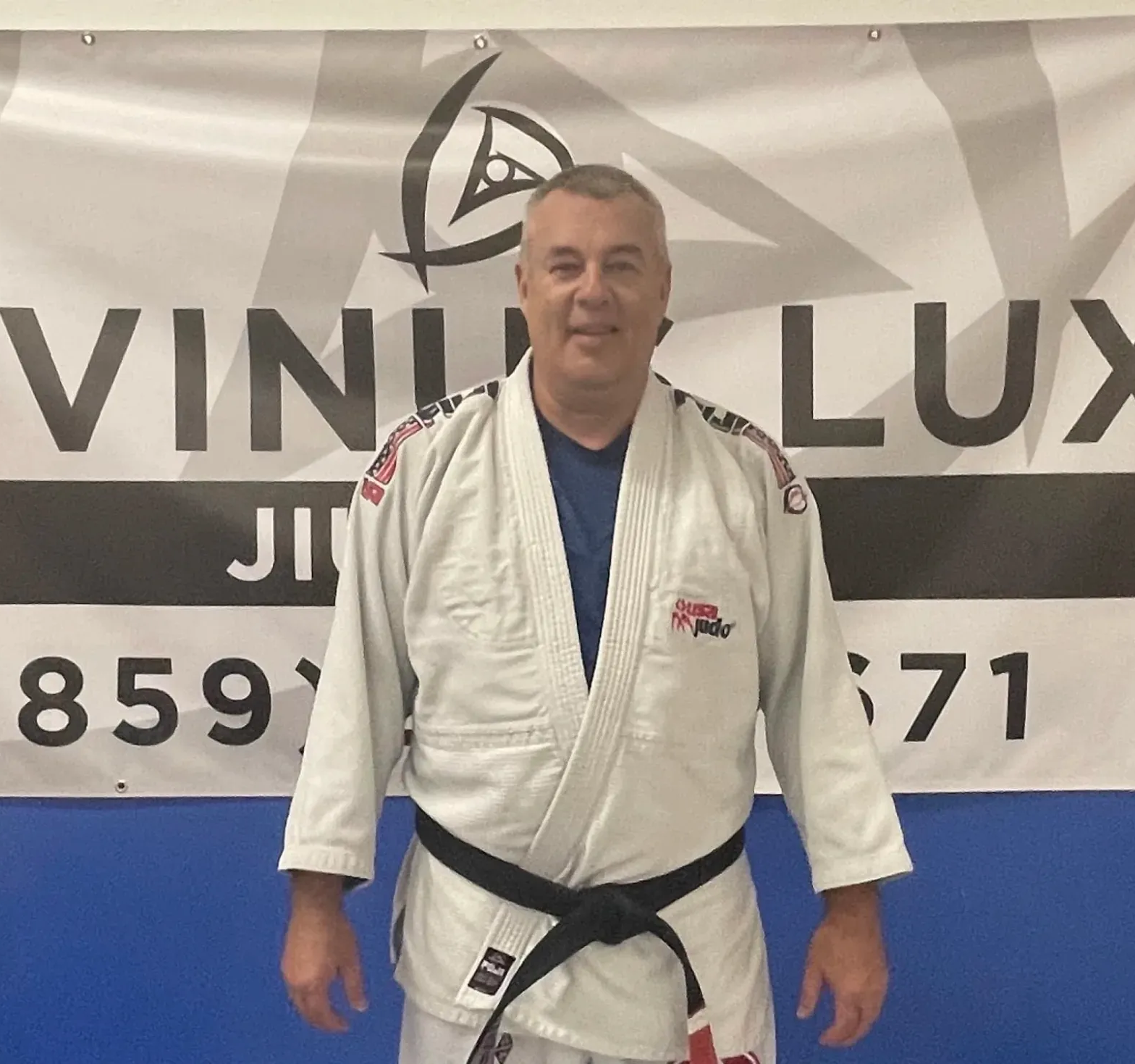 Man in a white jiu-jitsu gi with black belt standing before a Vini Lux Jiu-Jitsu banner
