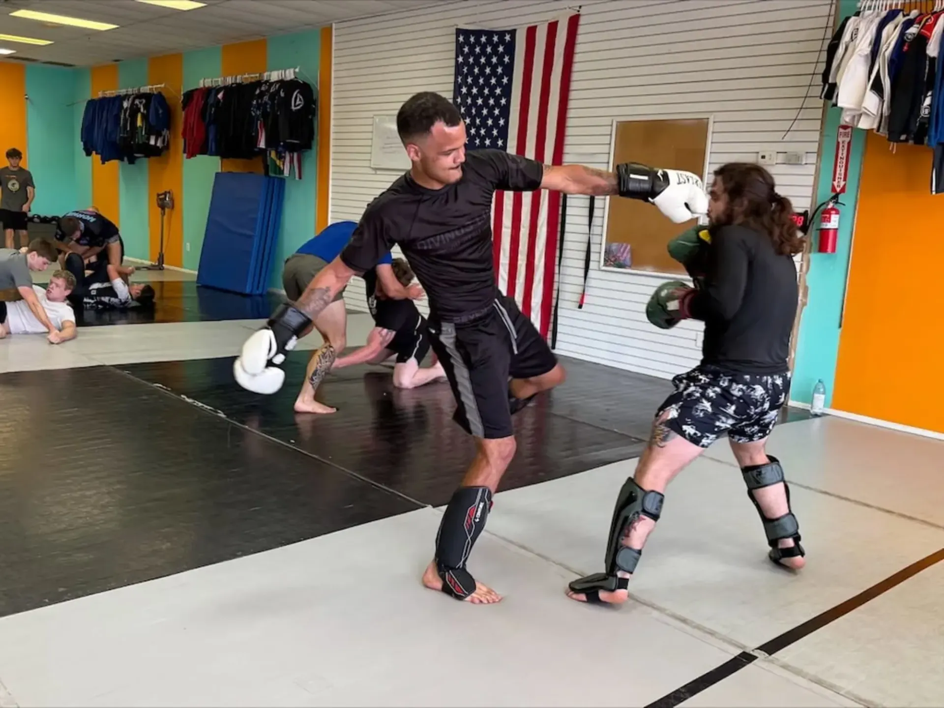 Two martial artists sparring with pads in a training gym, with gear and an American flag on the wall.