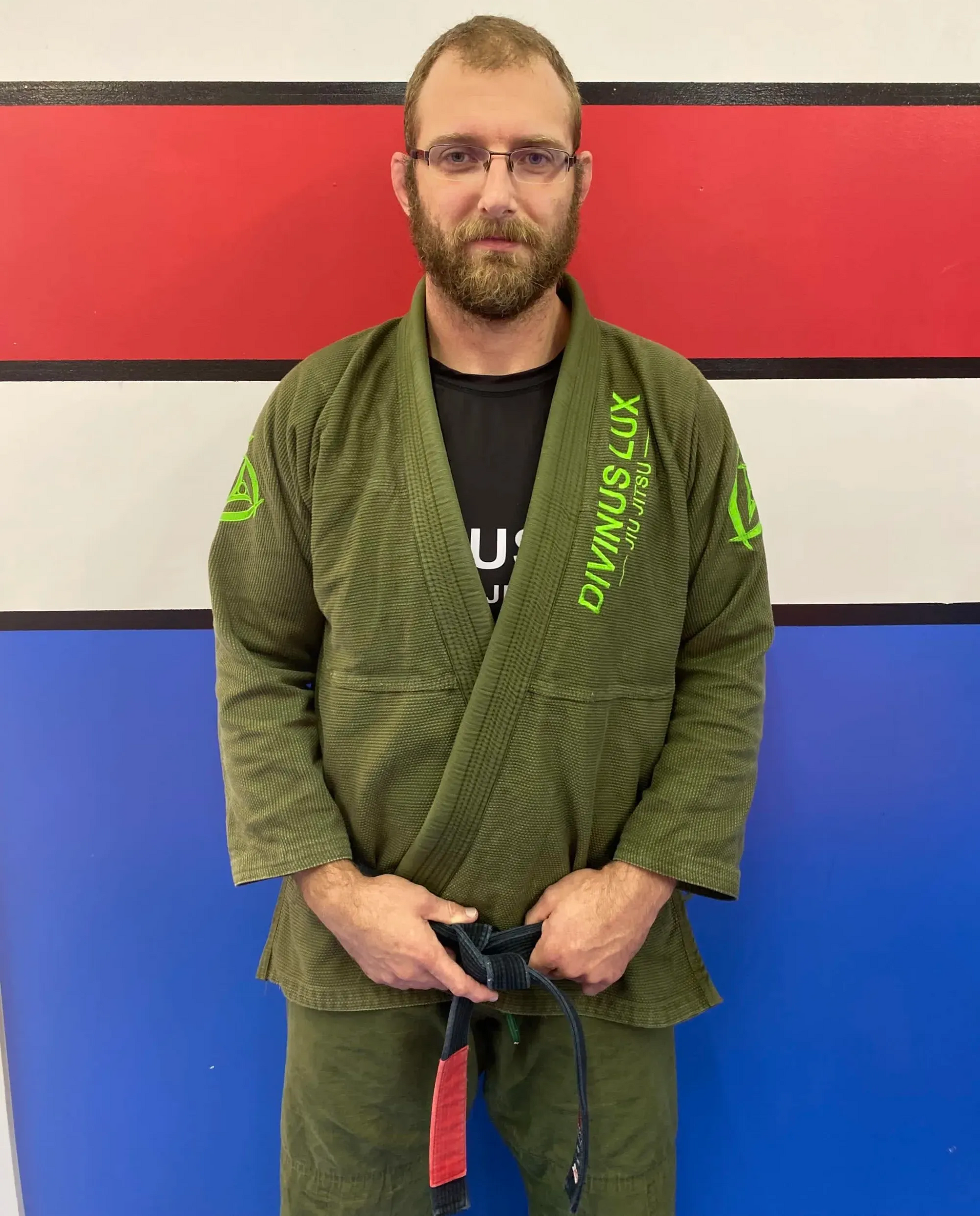 Man in green gi holding a belt, standing against a red, white, and blue wall.