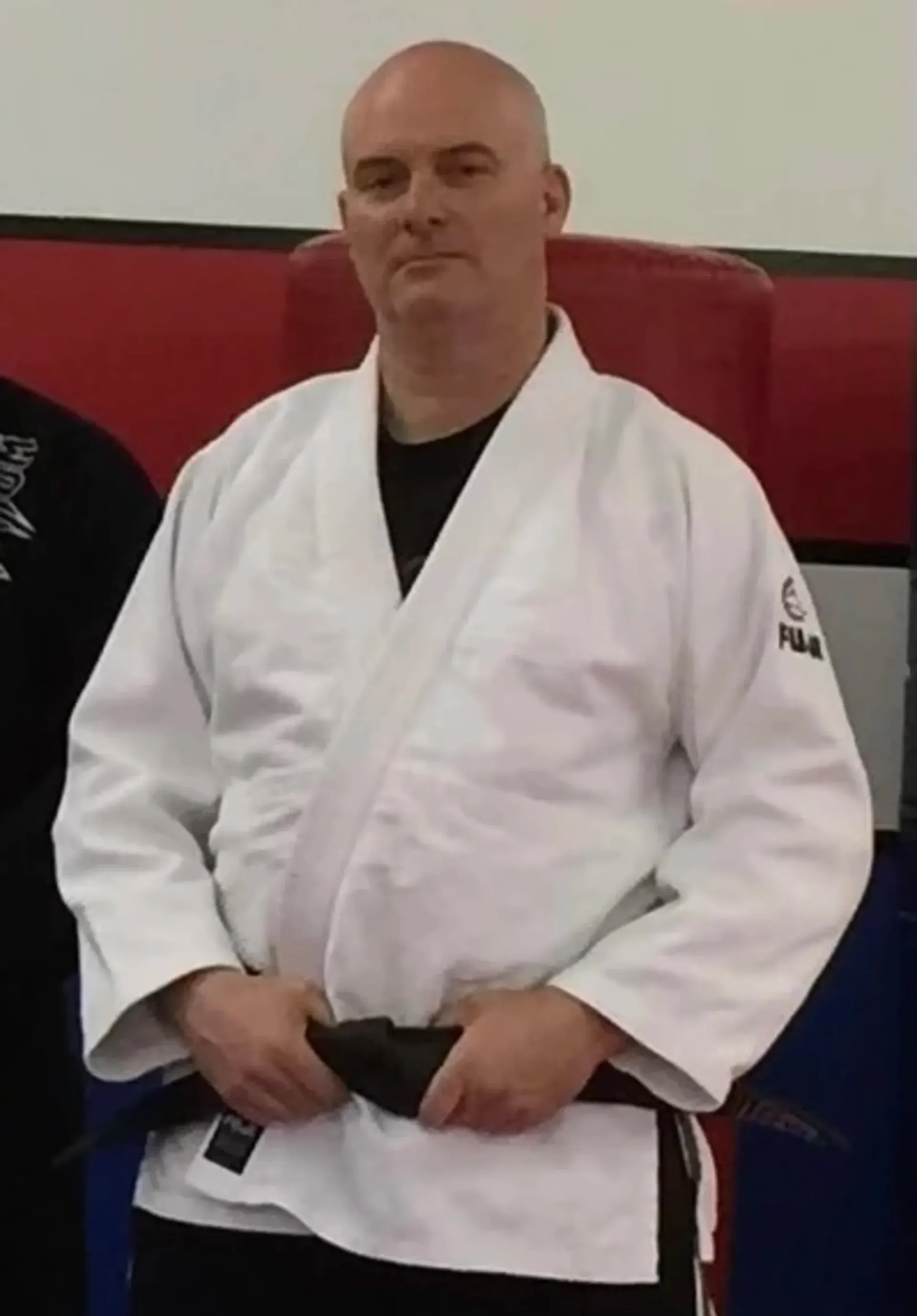 Martial artist in white gi with black belt standing indoors against a red and blue wall