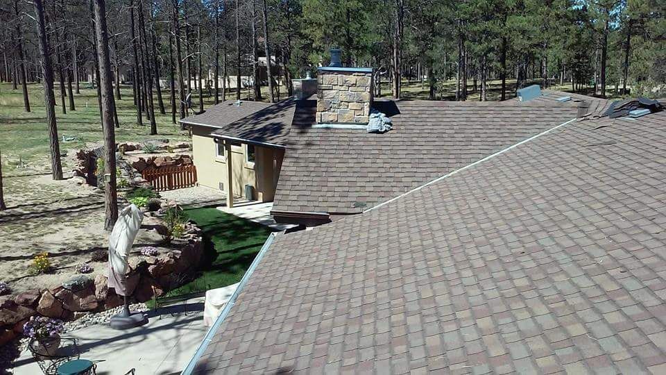A residential roof with brown shingles, a chimney, and trees in the background.