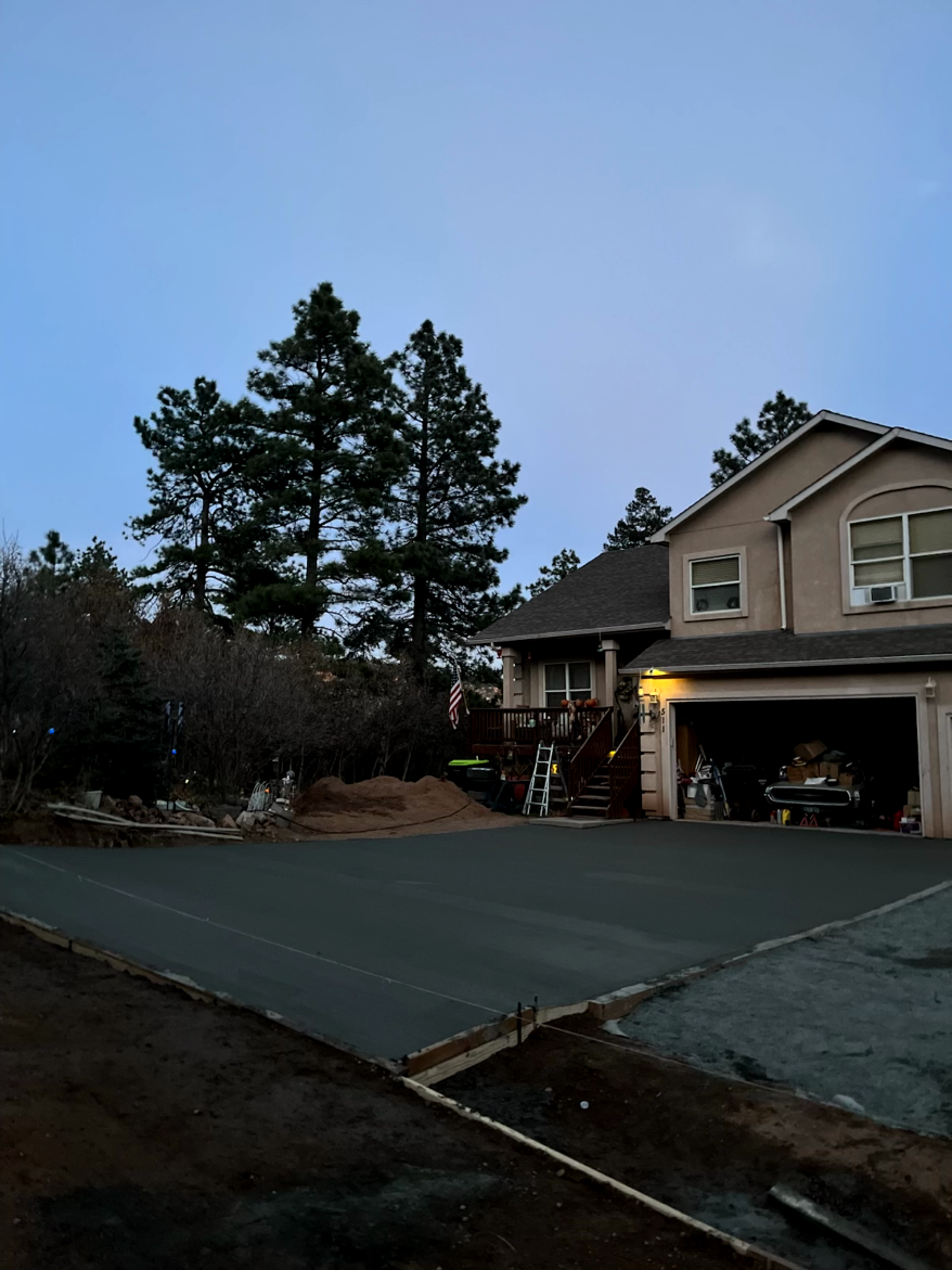 Newly paved driveway in front of a two-story beige house with an open garage, trees in background, dusk.