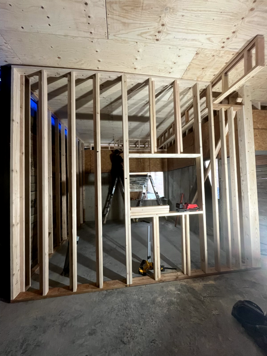 Interior wall framing under construction, with a window opening and a person on a ladder in the background.
